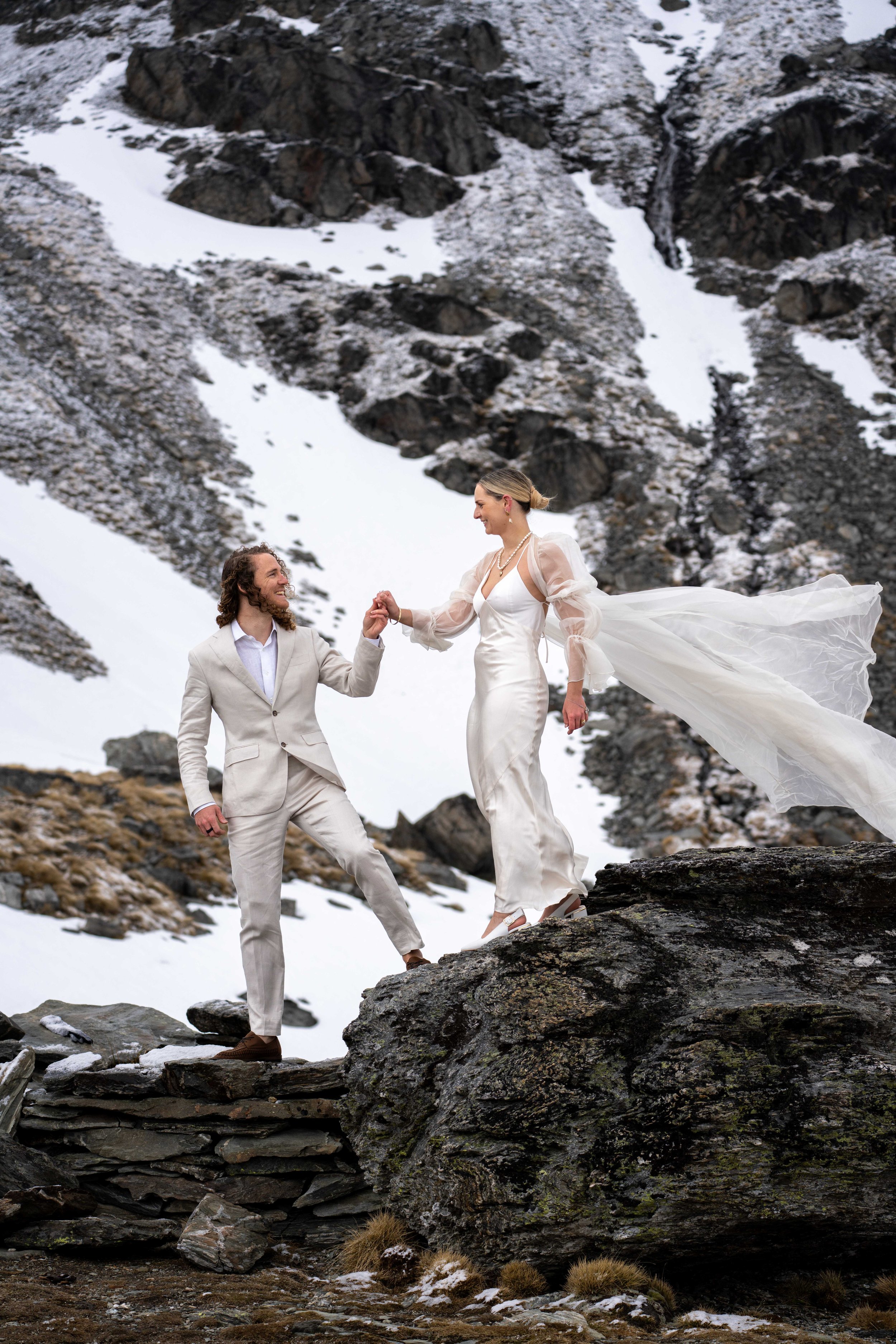 A couple in wedding attire standing on rocks in a snowy mountain landscape, holding hands and smiling at each other.