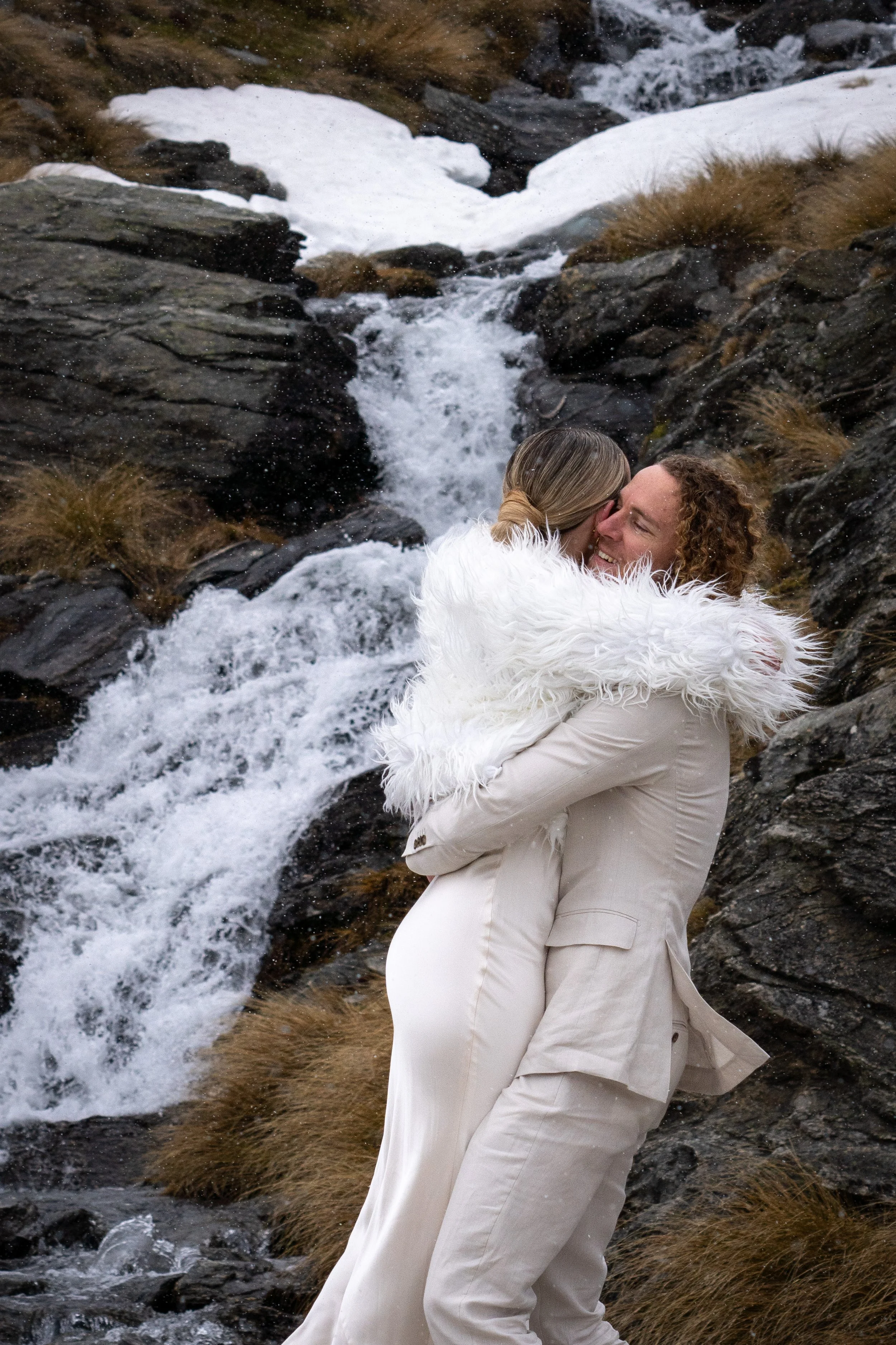 A couple hugging near a flowing mountain stream, with snow patches and dry grass in the background.
