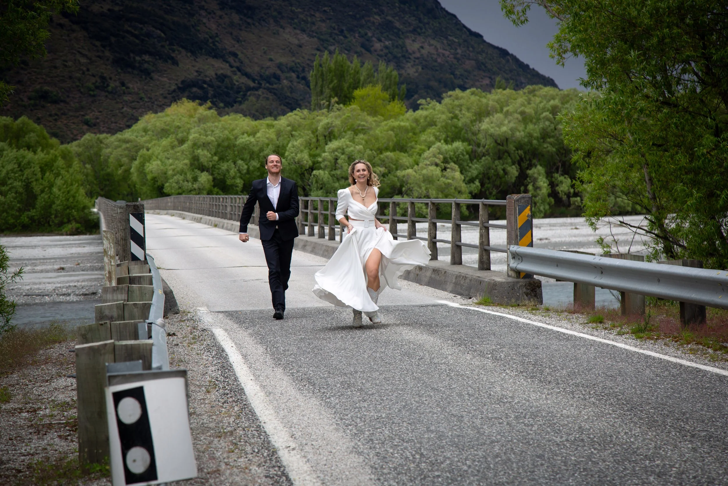 A joyful bride in a white wedding dress and a groom in a black suit running across a small bridge over a river, surrounded by green trees and mountains.
