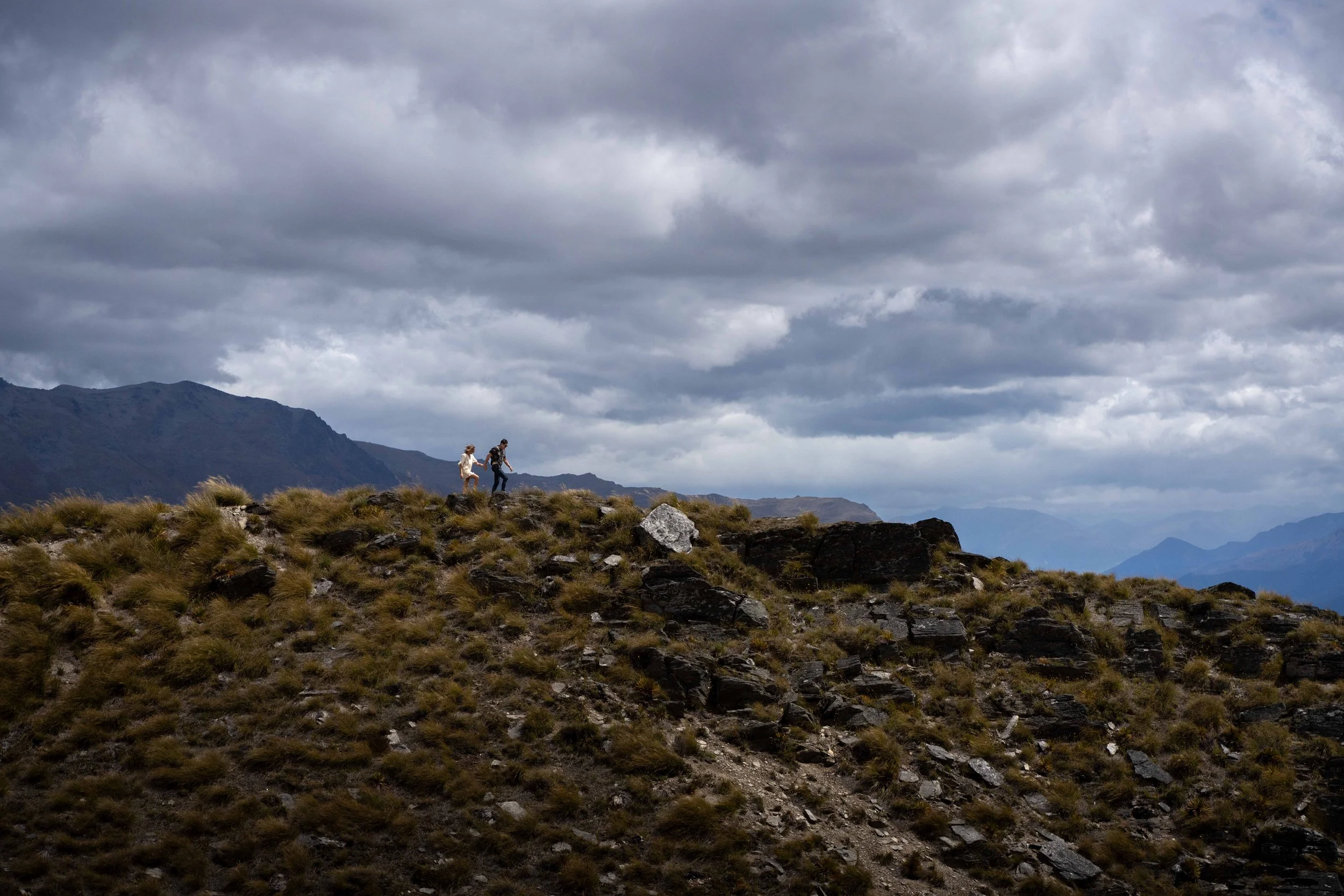 Two hikers walking along a rocky, grassy hillside under a cloudy sky with mountains in the distance.