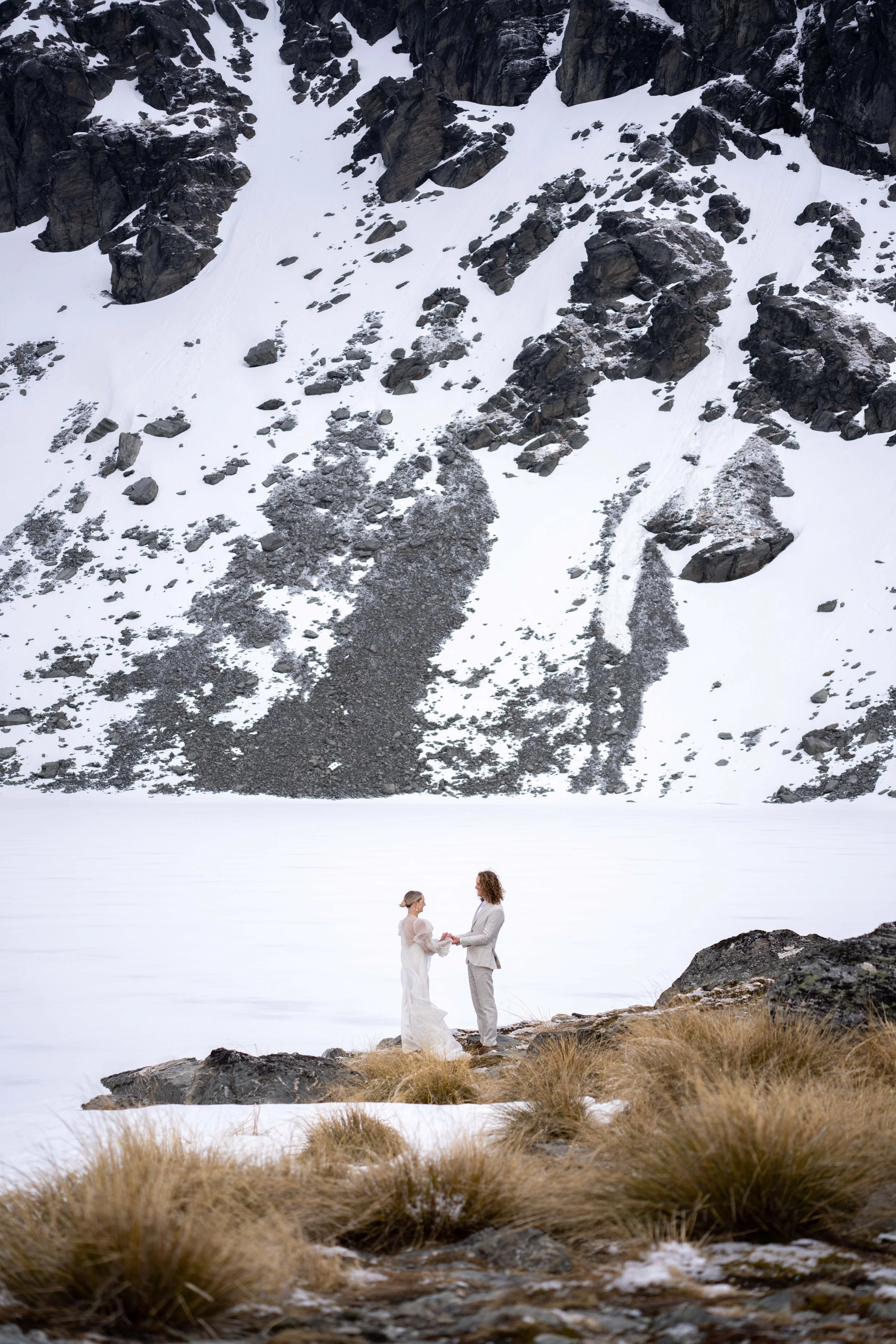 A couple on their wedding day standing outdoors near a rocky, snowy landscape with a mountain in the background, holding hands.