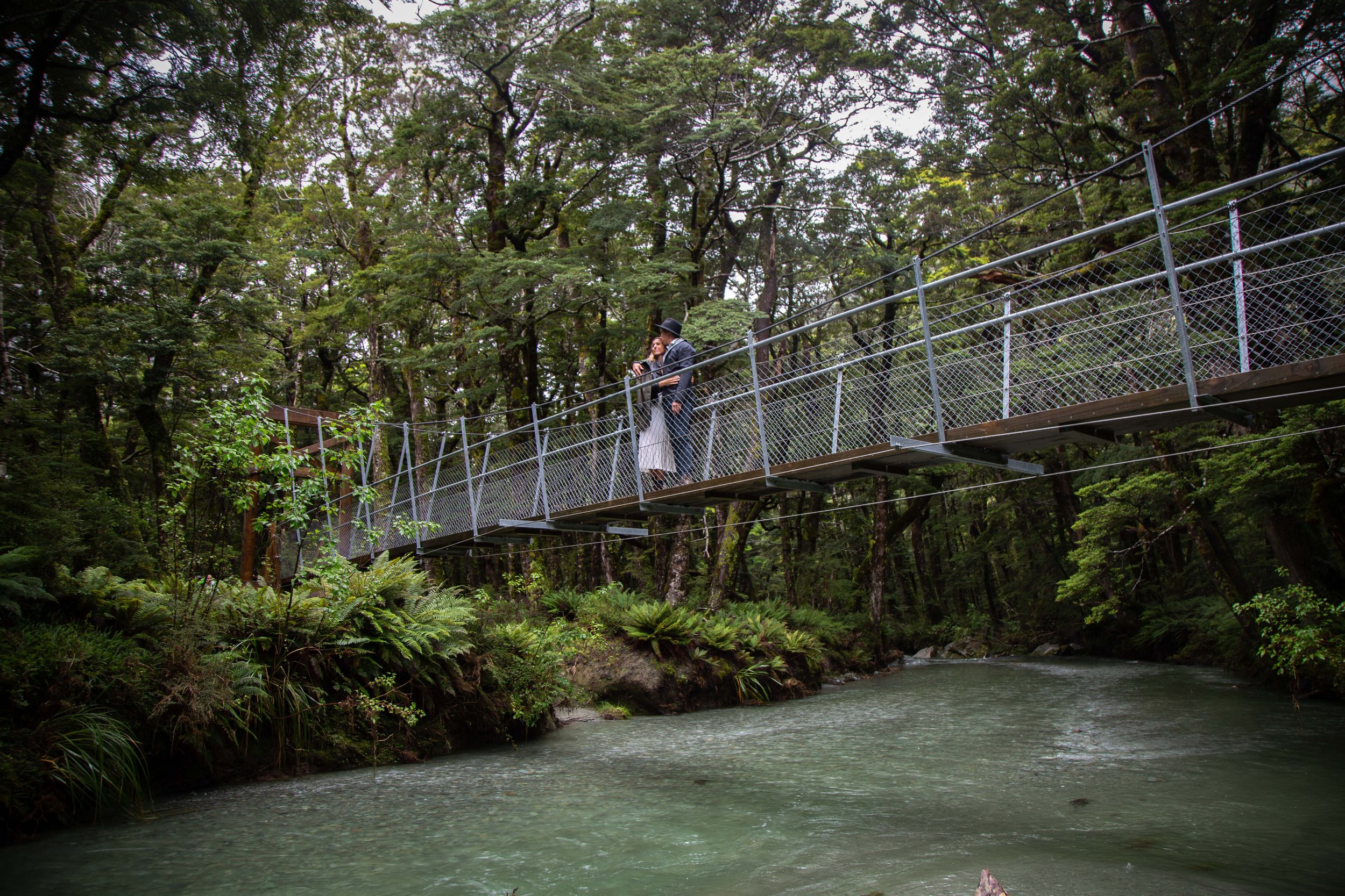 A couple in formal attire standing on a suspension bridge over a river in a forested area.