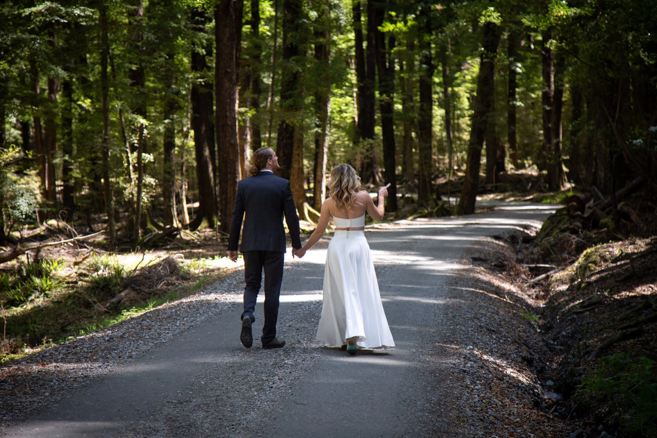 A couple in wedding attire walking hand in hand on a forest trail, holding hands, with tall trees and dappled sunlight.