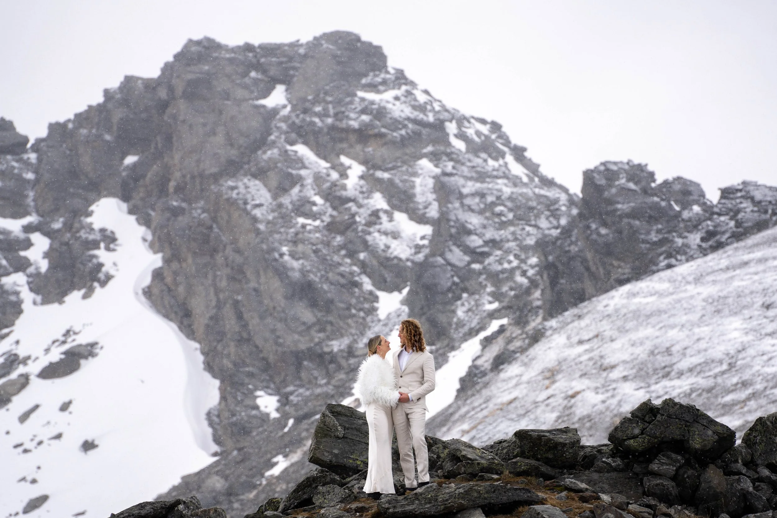 Two women in wedding attire standing on rocks in a snowy mountainous landscape, holding hands and gazing at each other.