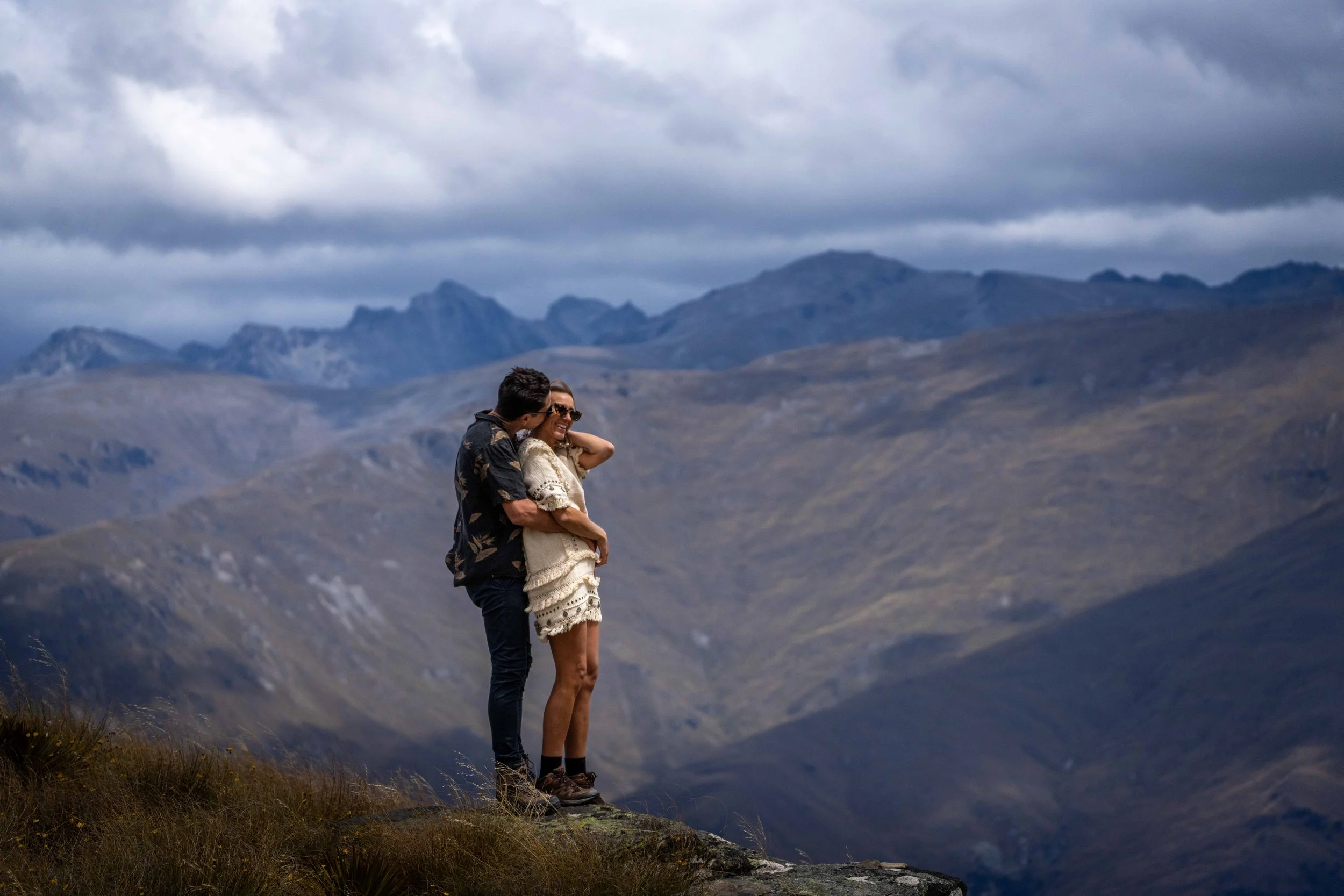 A couple standing on a rocky ledge in front of mountain ranges and cloudy sky, sharing a romantic moment.