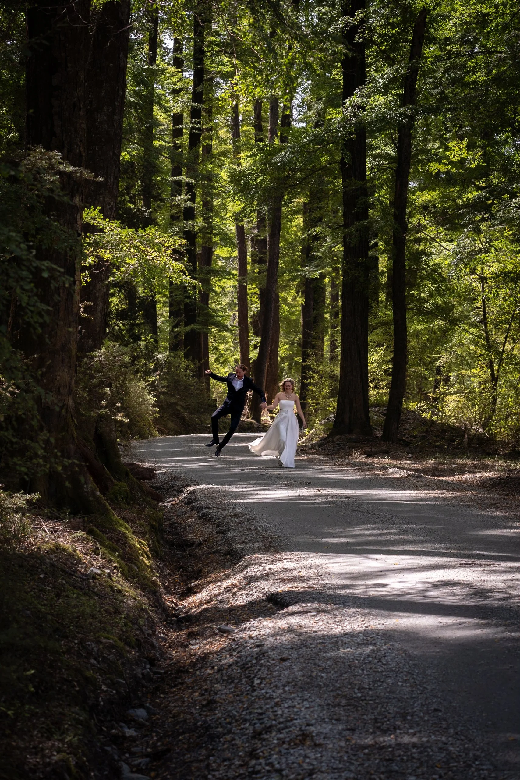 A bride and groom holding hands and jumping on a forest trail surrounded by tall trees and lush green foliage.