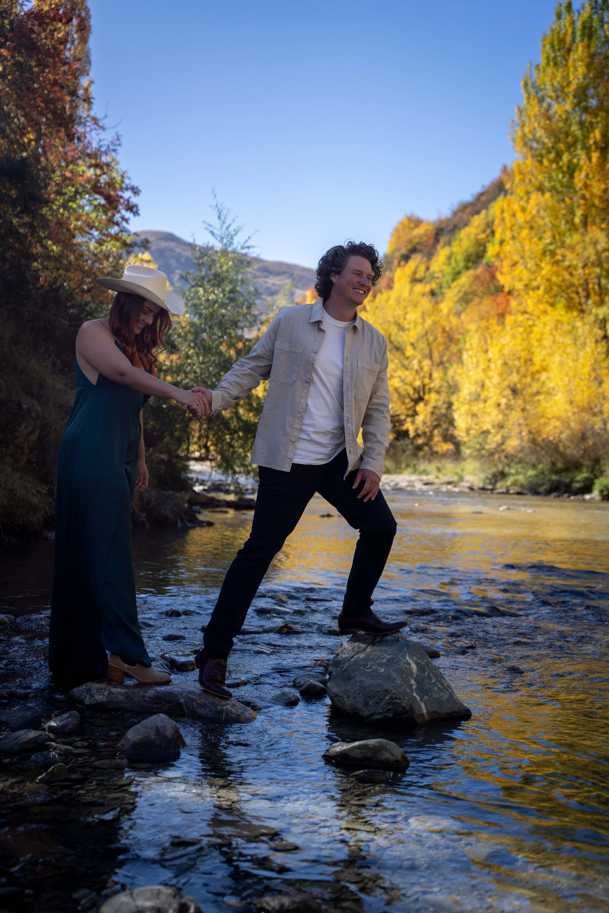 A couple holding hands and walking on rocks in a shallow river in a forested area during autumn.