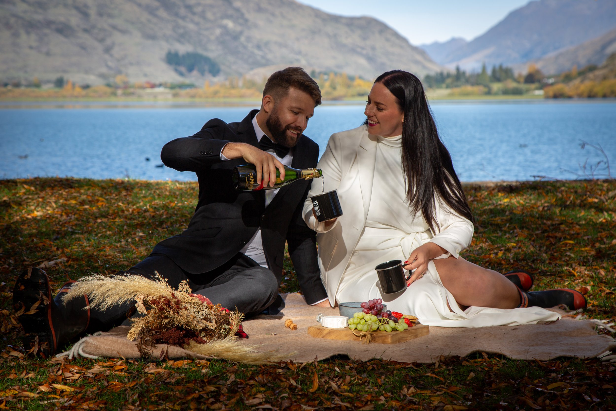 A couple in formal attire having a picnic outdoors by a lake, with mountains in the background. They are sitting on a blanket, with fruit and drinks, and smiling as the man pours a drink into the woman's mug.