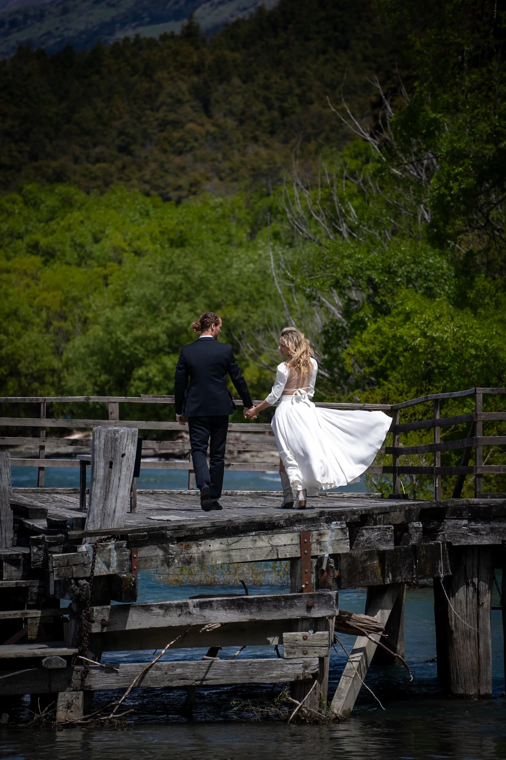 A couple dressed in wedding attire holding hands while walking on a rustic wooden dock over water, with lush green trees and mountains in the background.