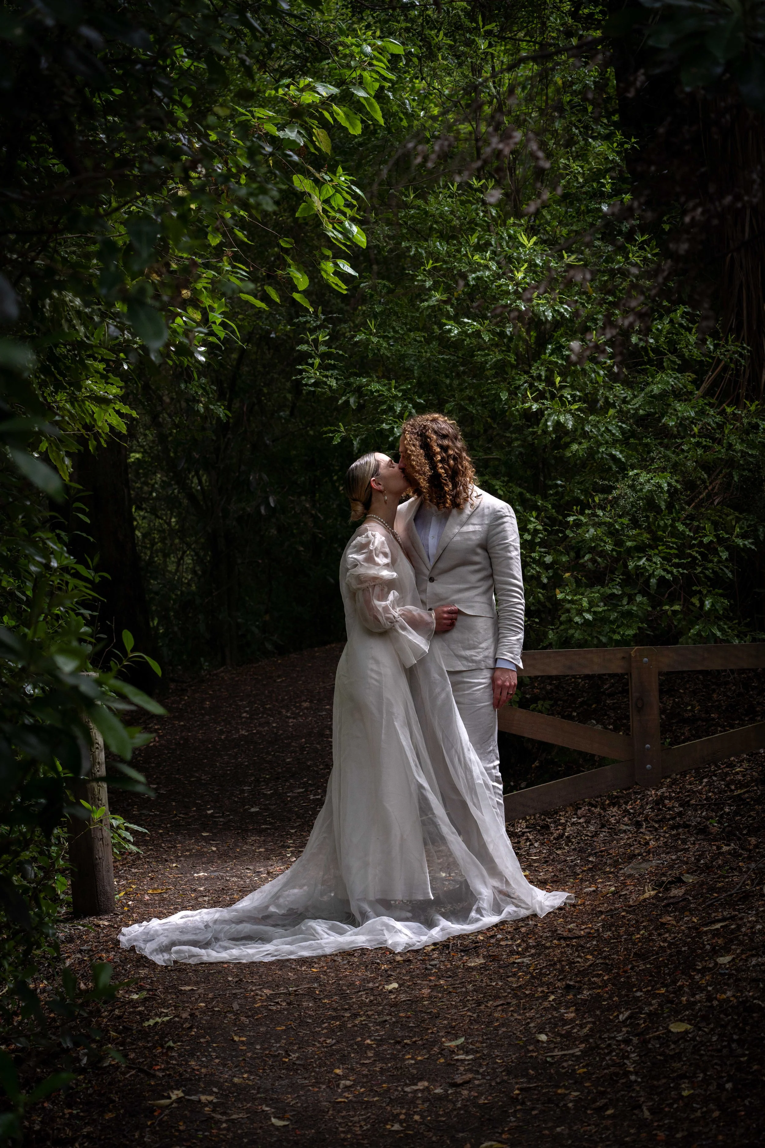 A couple dressed in white wedding attire sharing a kiss on a wooded trail surrounded by green foliage.
