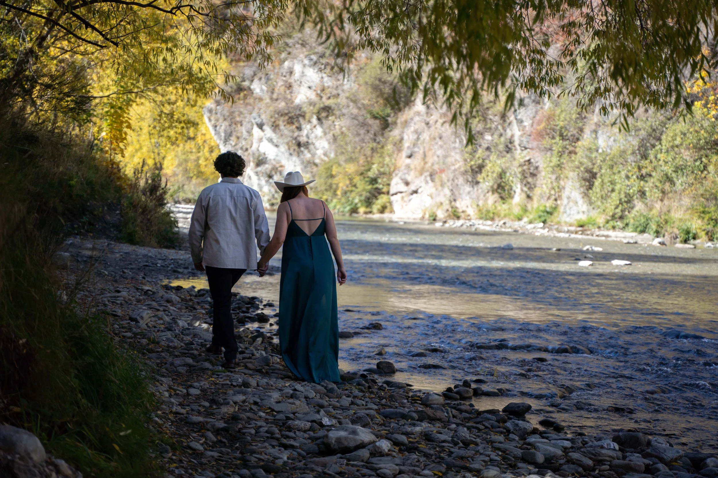 A couple walking hand in hand along a rocky riverside surrounded by trees and mountains.