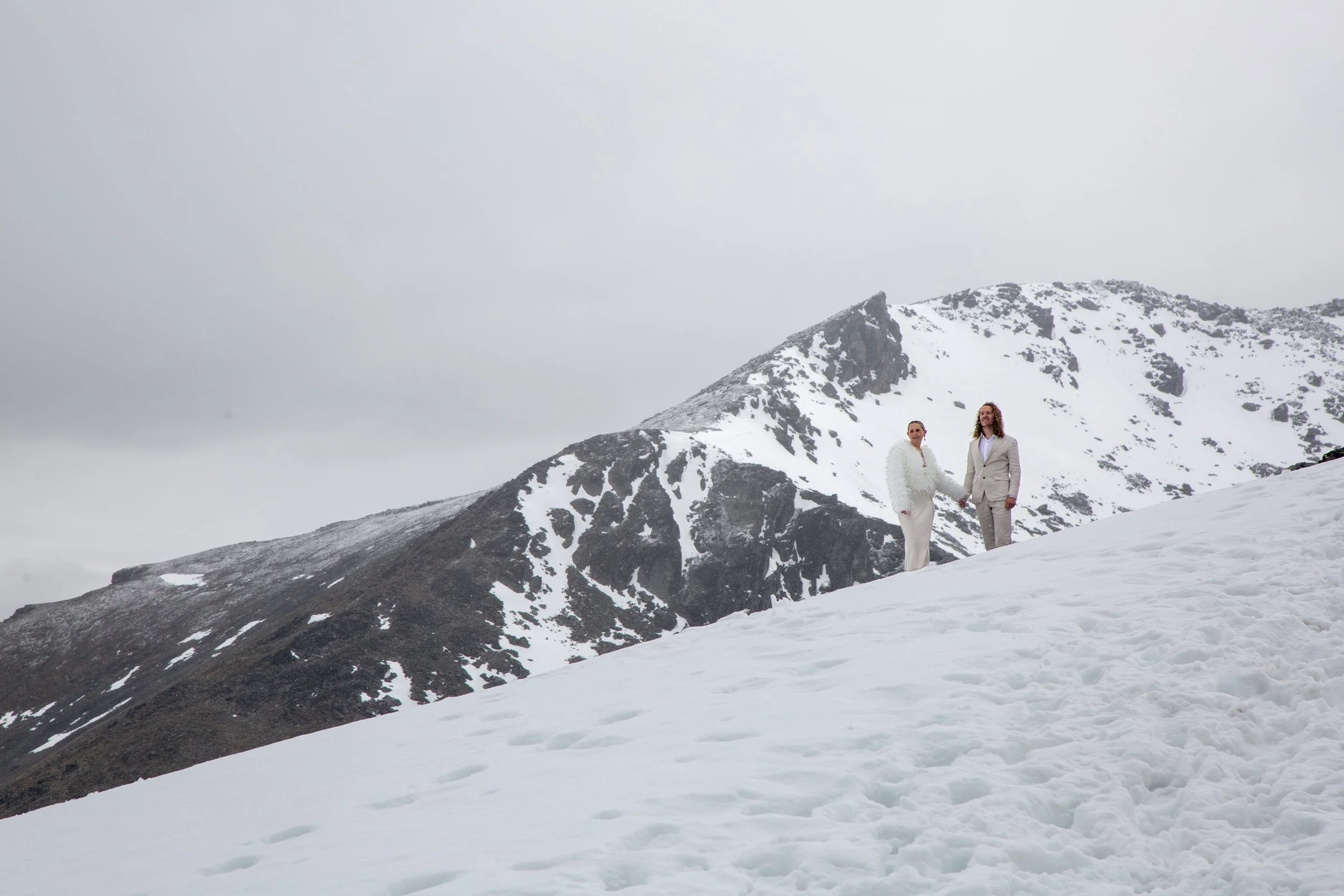 Two women in white and gray suits hold hands on a snowy mountain slope with a snow-covered mountain and cloudy sky in the background.