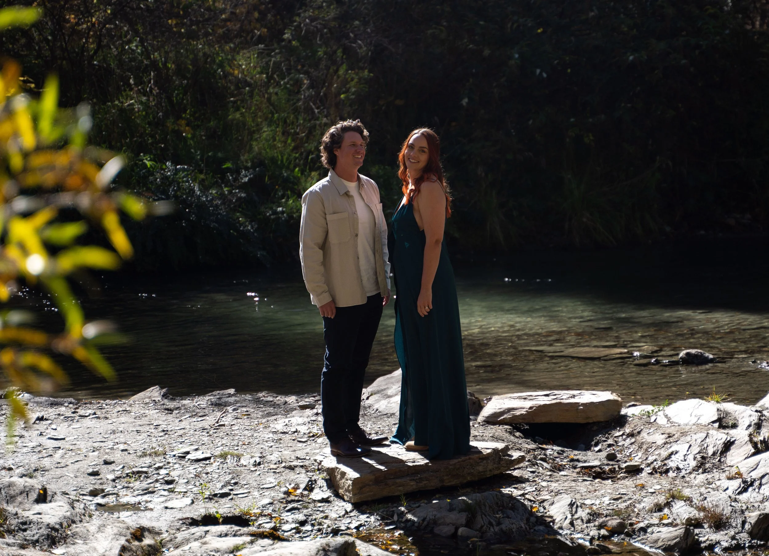 A man and woman standing on a large flat rock by a creek surrounded by trees, smiling at each other during daytime.