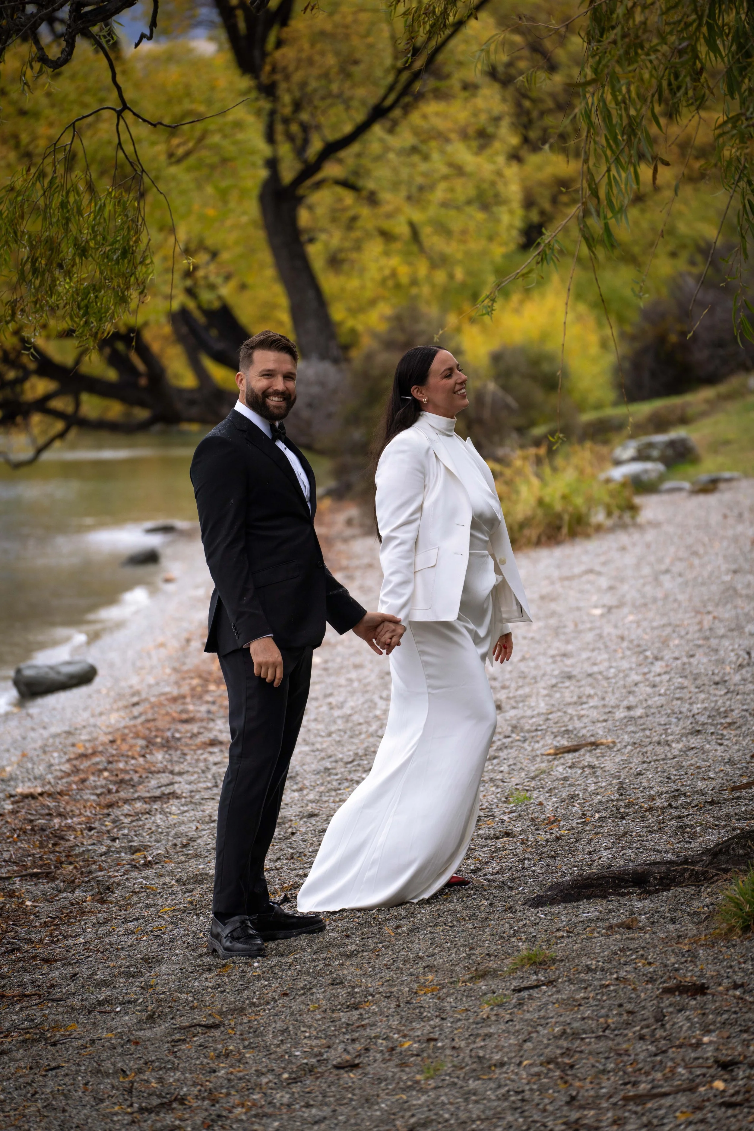 A newlywed couple holding hands by a lake during fall, with trees showing yellow leaves, standing on a gravel shore.