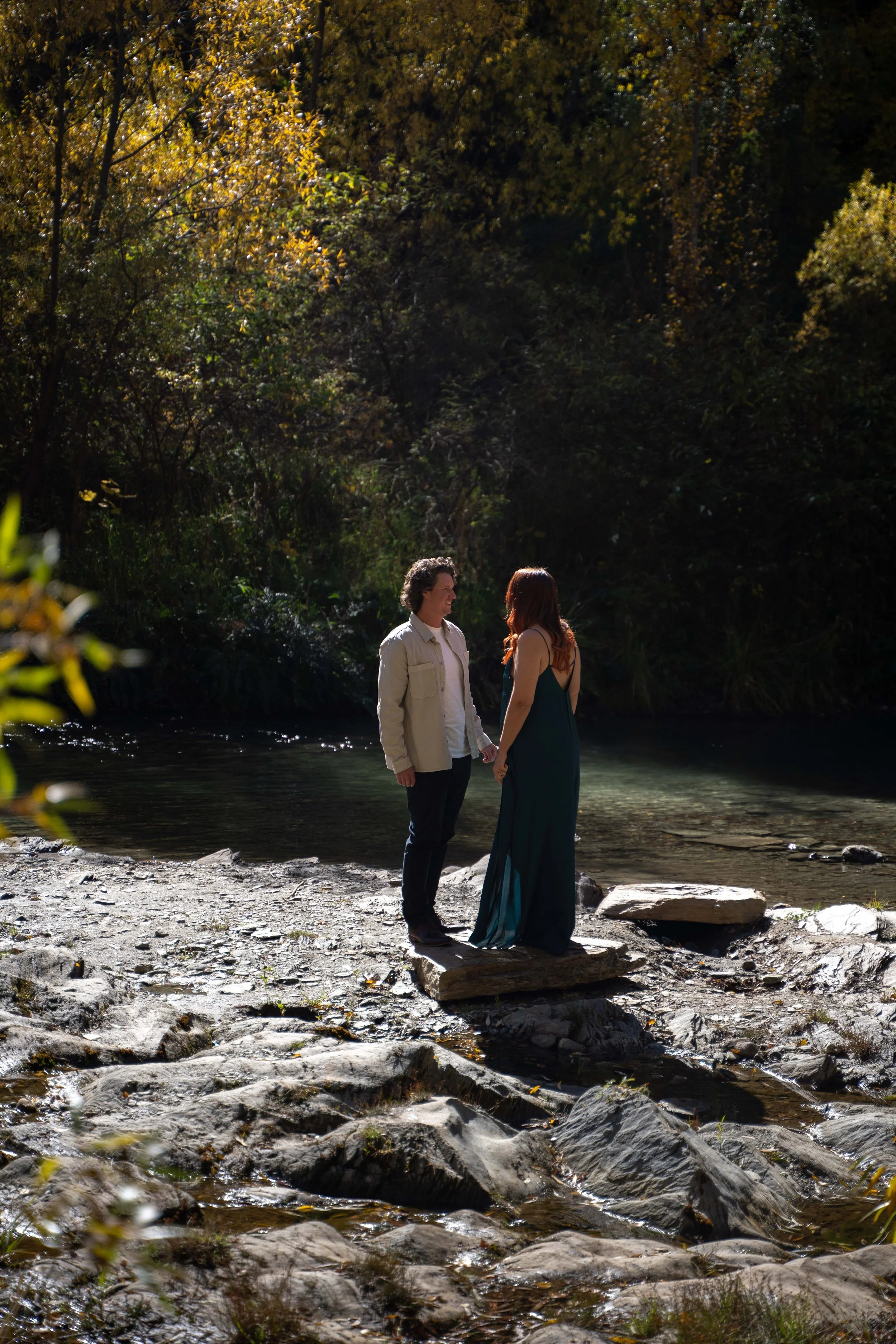 A couple standing on a rocky stream in a forest, gazing at each other and holding hands, illuminated by sunlight filtering through trees.