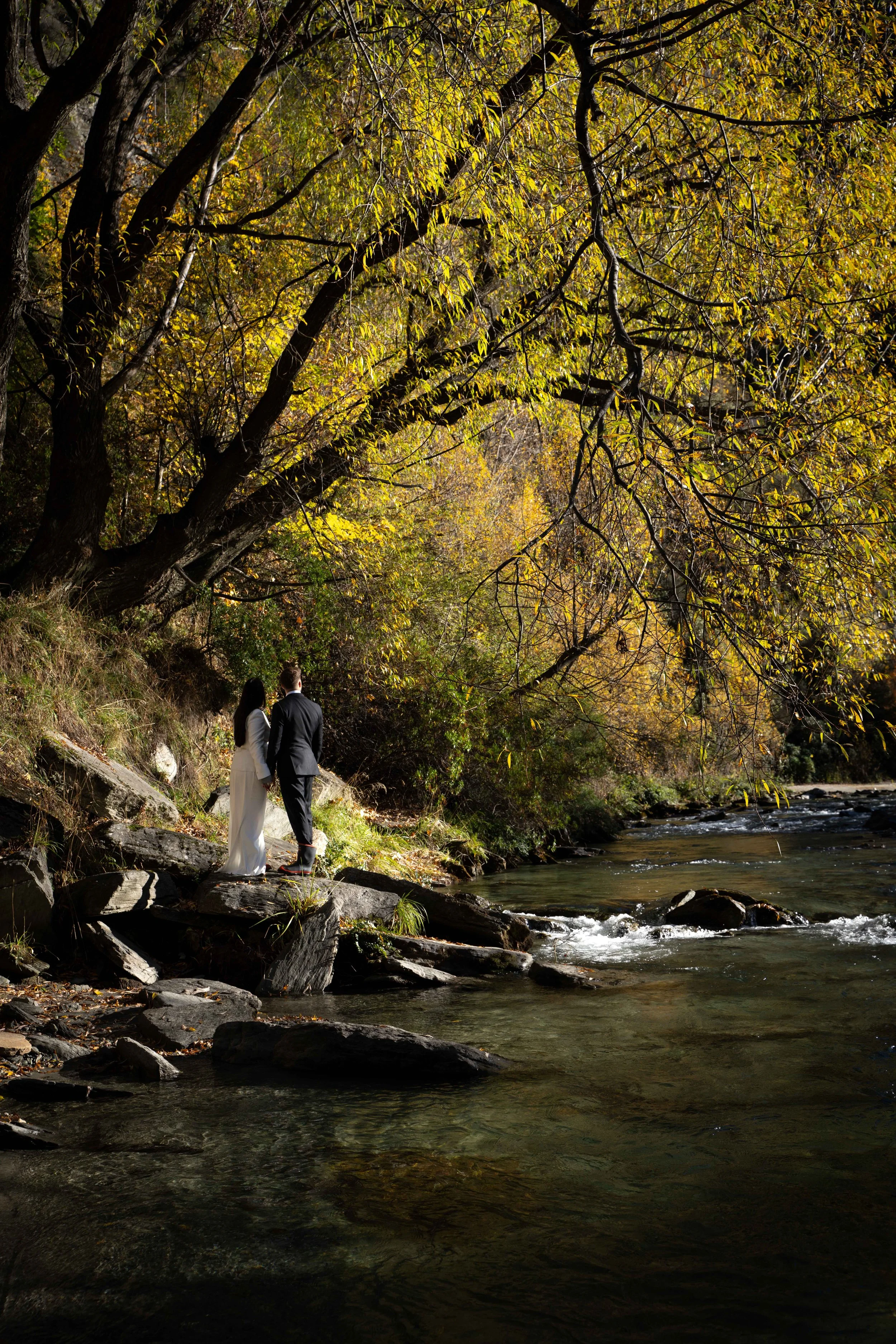 A couple in wedding attire holding hands on rocks beside a flowing river in a forest with autumn-colored trees.