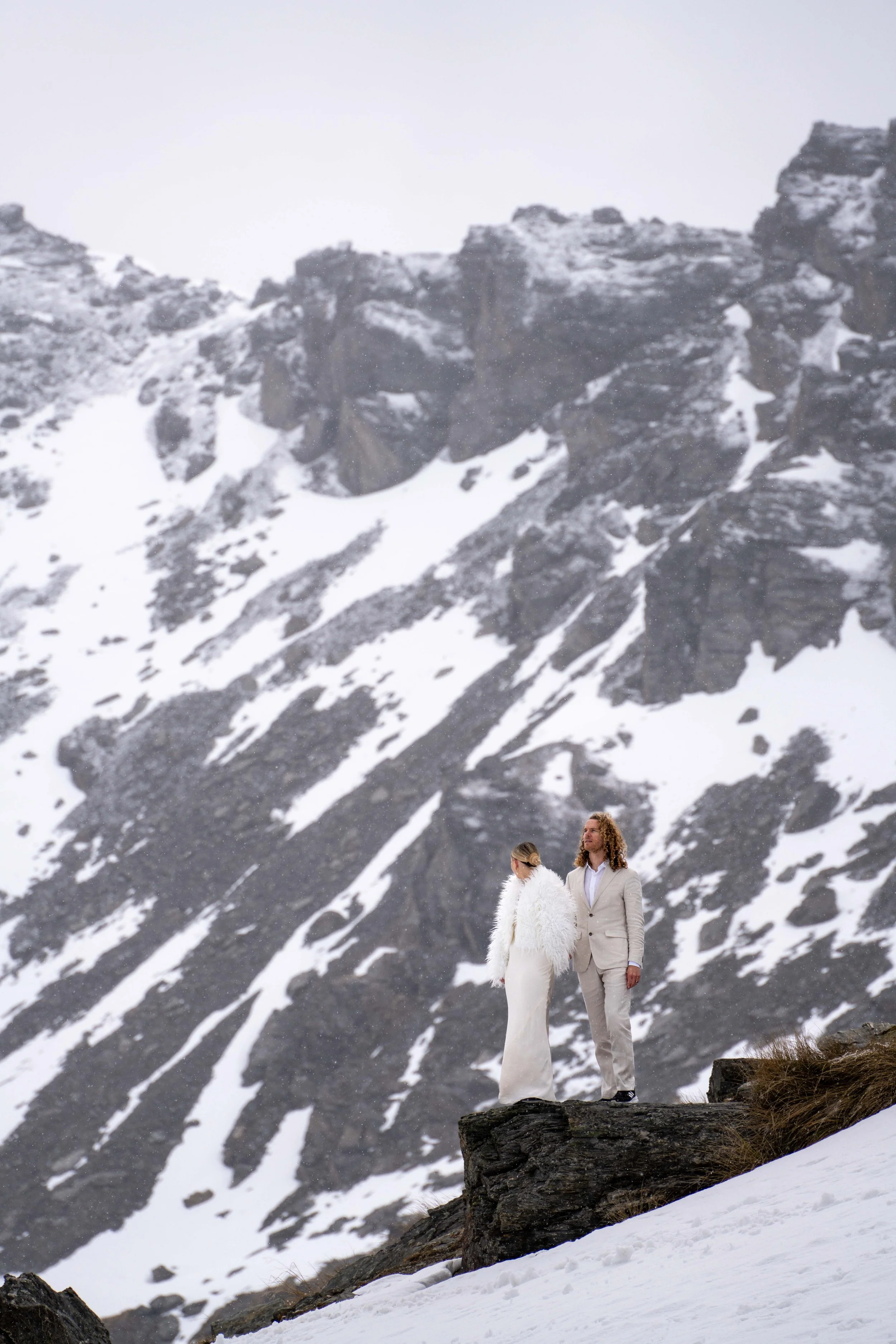 A couple in white wedding attire stands on a rocky outcrop in a snowy mountainous landscape with steep, rugged cliffs in the background.