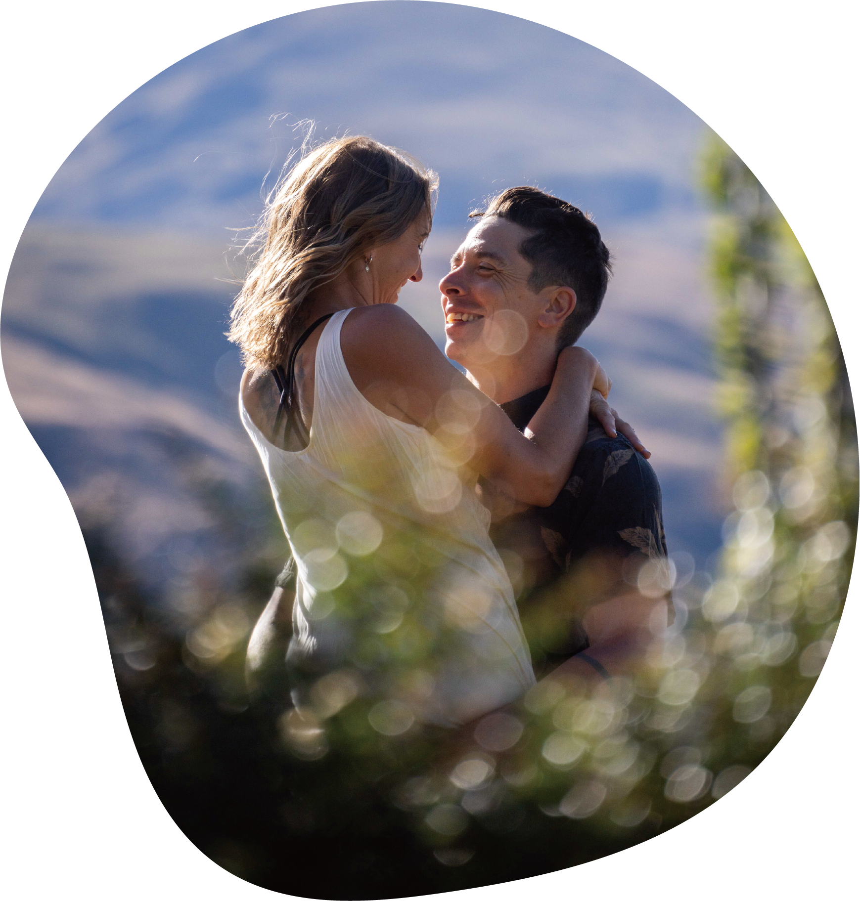 A couple smiling and embracing outdoors with mountains and blue sky in the background, and sunlight creating lens flare and bokeh effects.