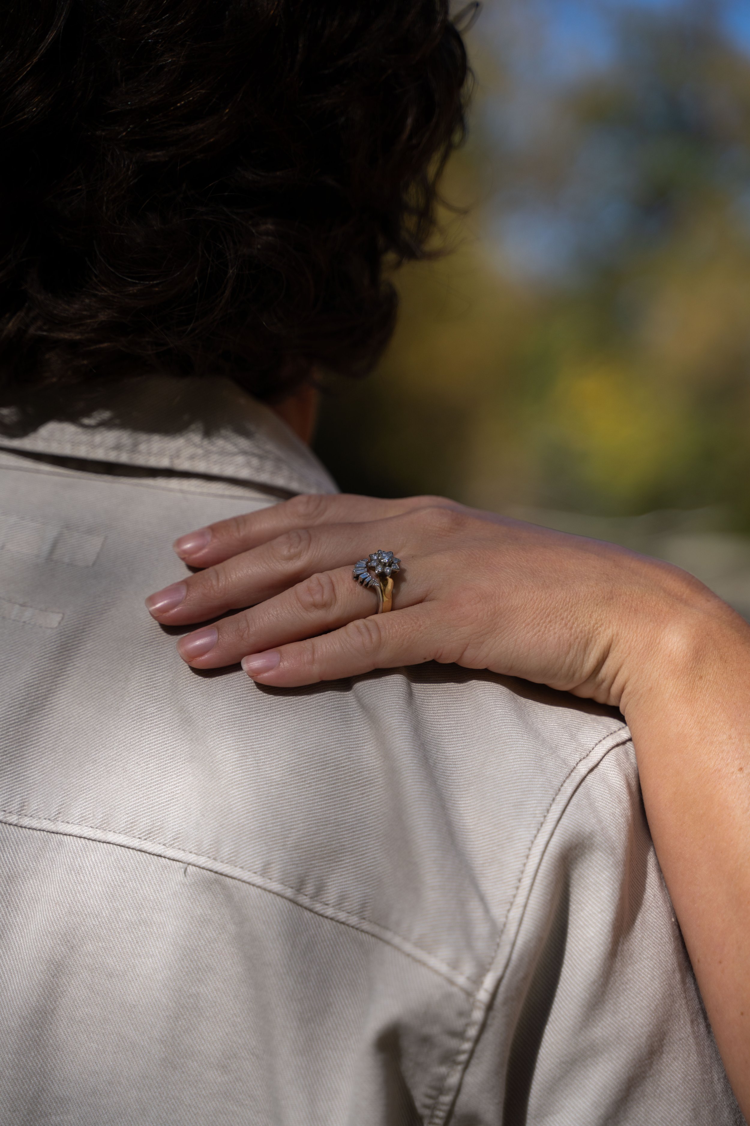 Close-up of a woman's hand resting on her shoulder, showcasing a floral ring with diamonds on her finger, against a background of blurred outdoor scenery.