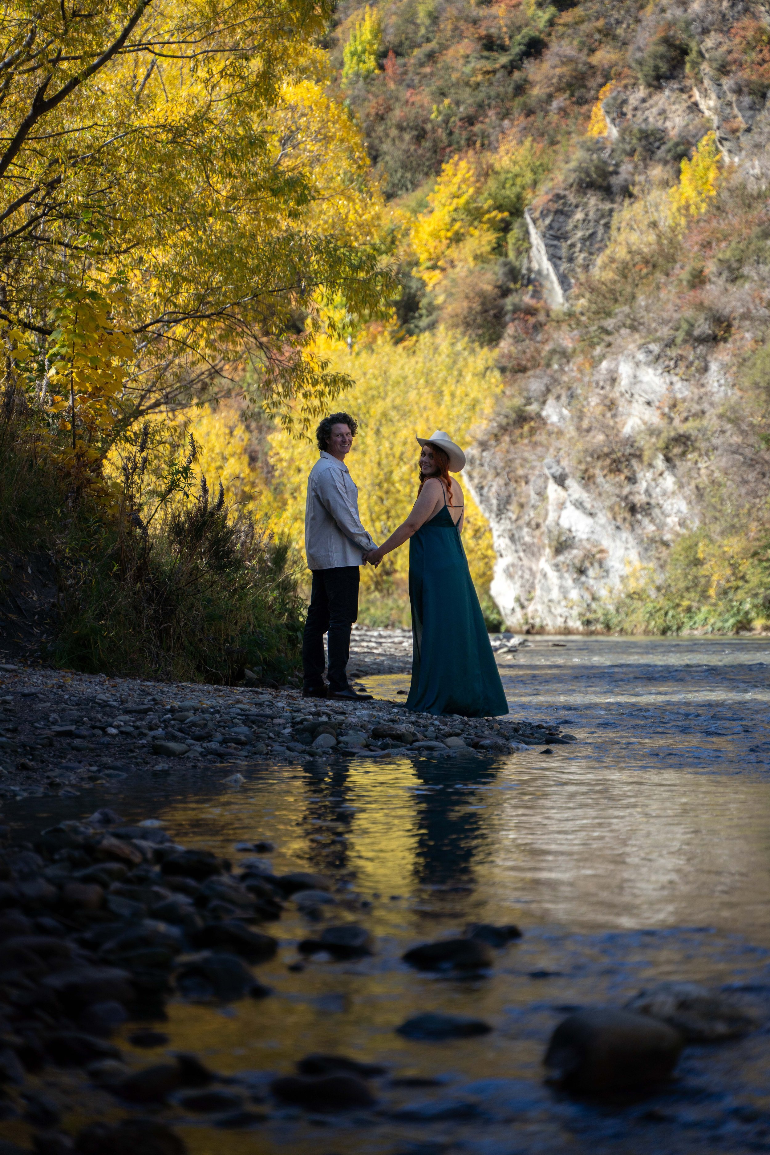 A couple holding hands standing by a river, surrounded by autumn-colored trees and rocky terrain, with mountains in the background.