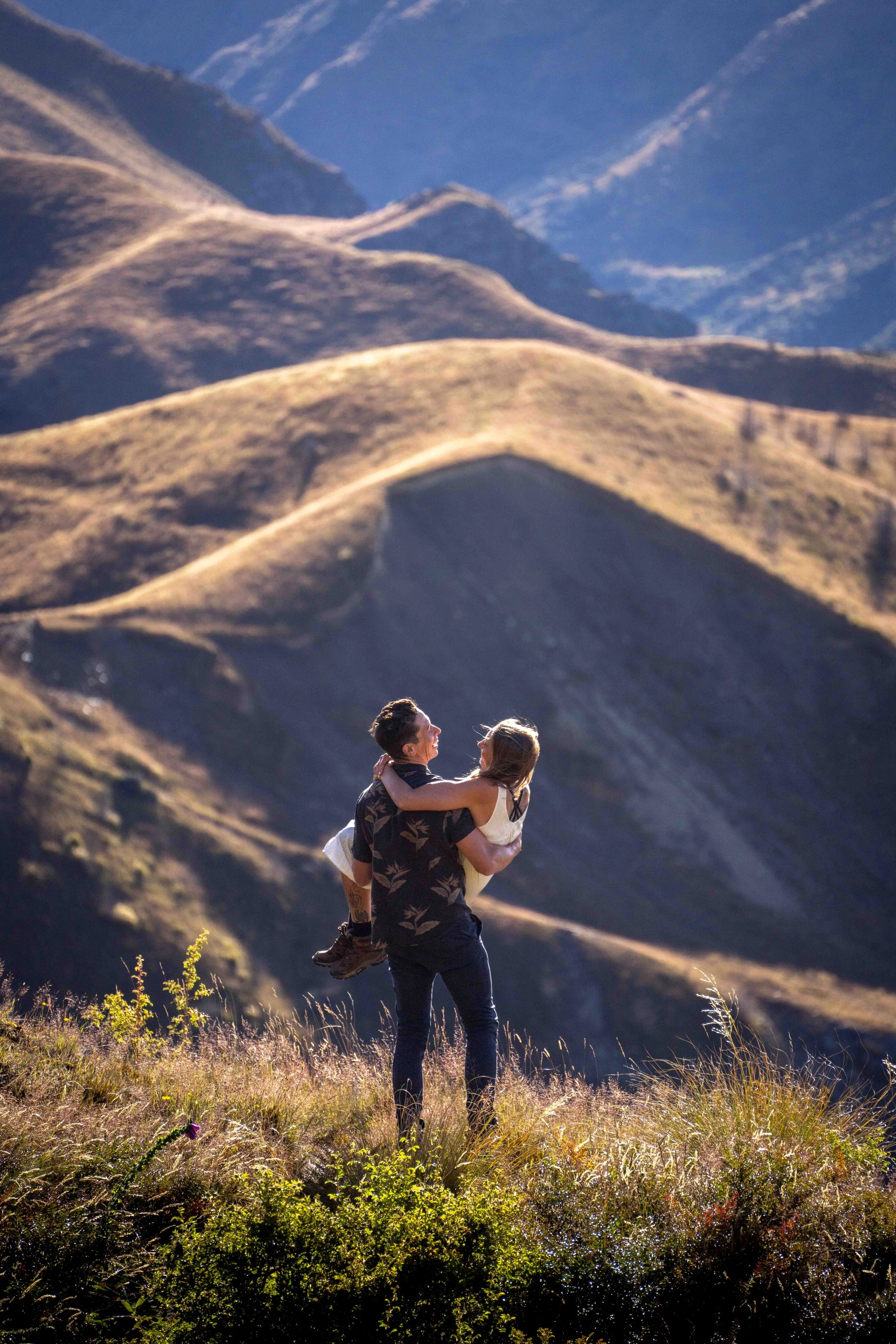 A man is holding a woman in his arms in a grassy field with rolling hills in the background.