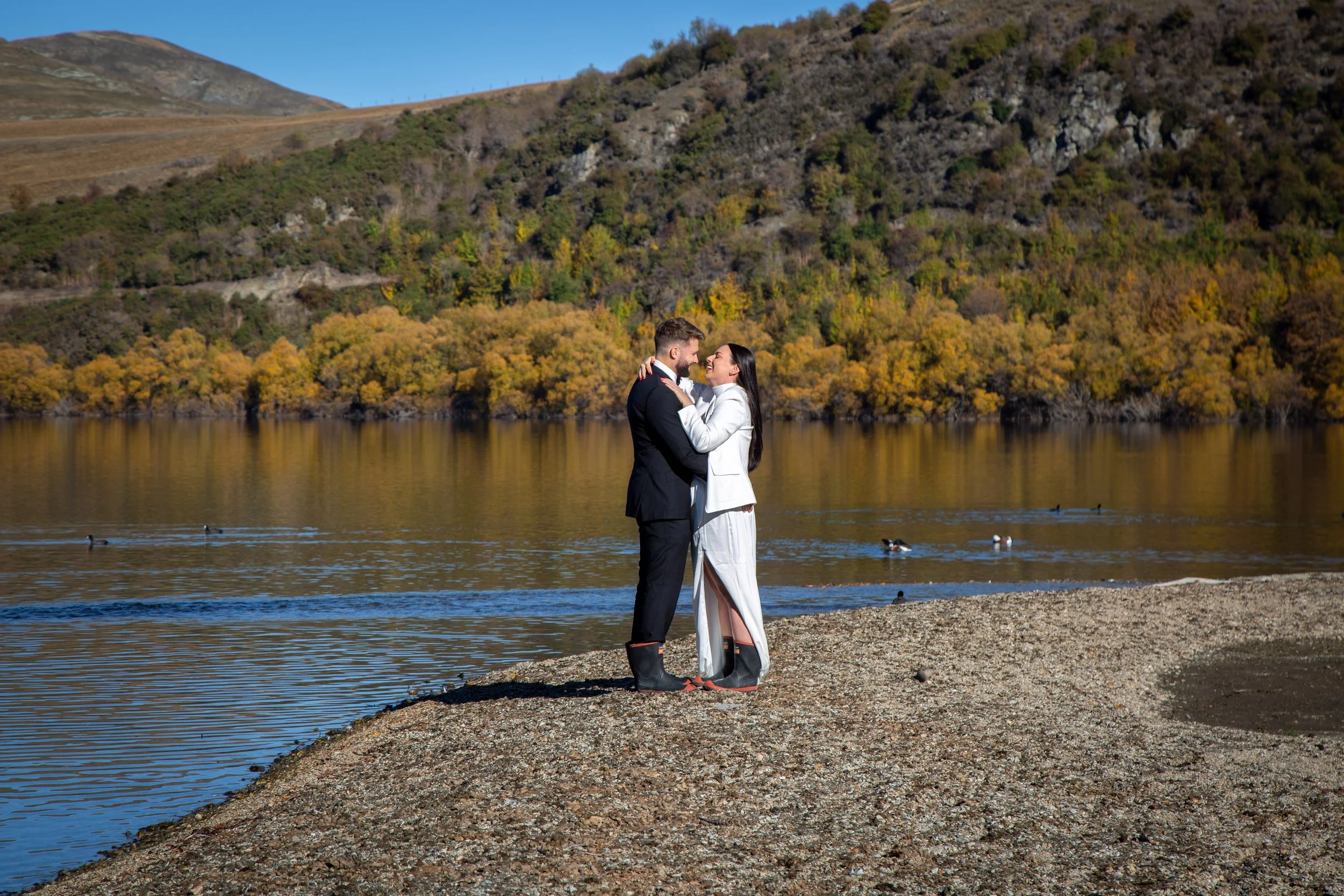 A couple dressed in wedding attire standing on a pebbled shore by a lake, embracing and smiling at each other, with autumn-colored trees and hills in the background.
