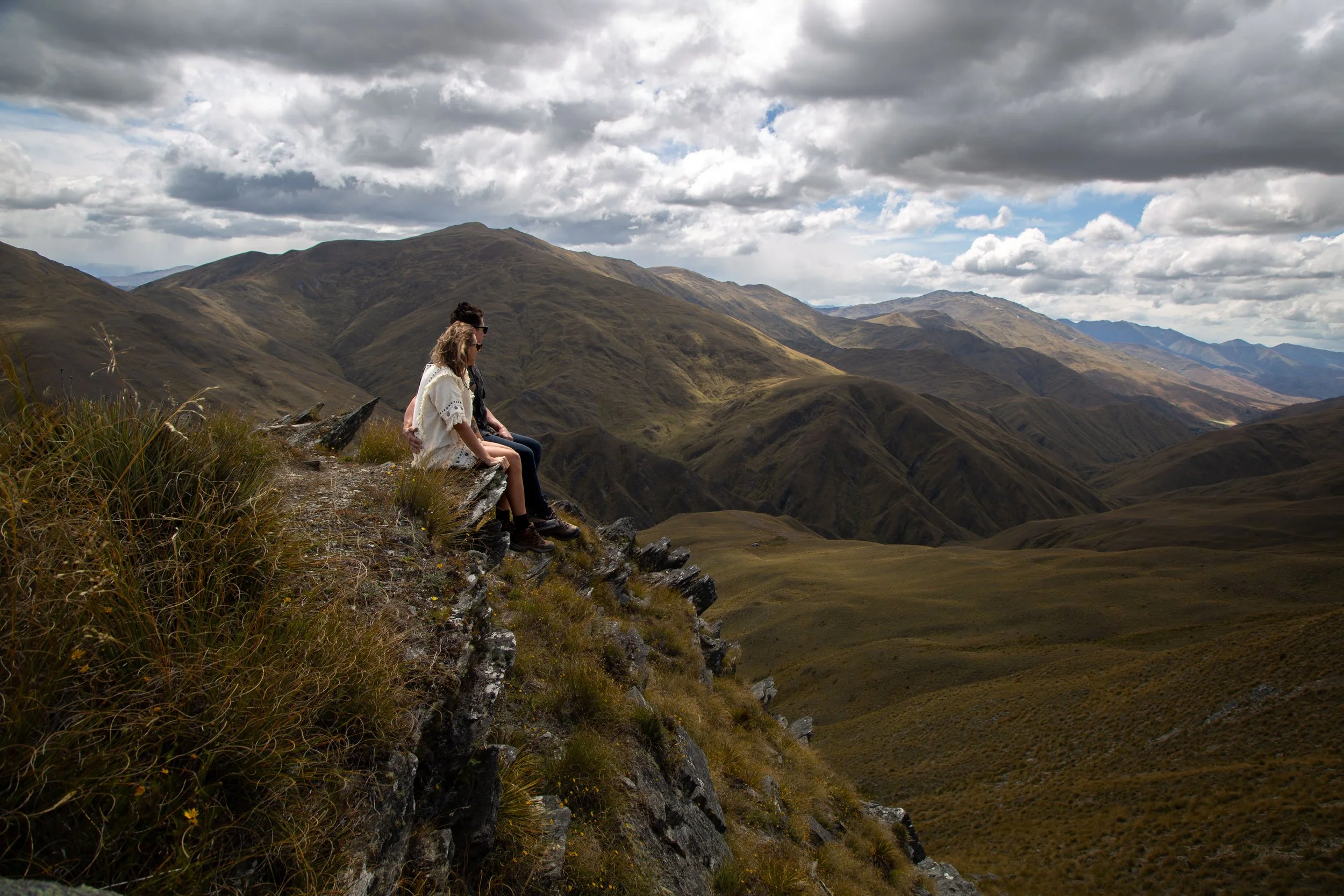 Three people sitting on a ledge overlooking a mountain valley, with cloudy sky above.