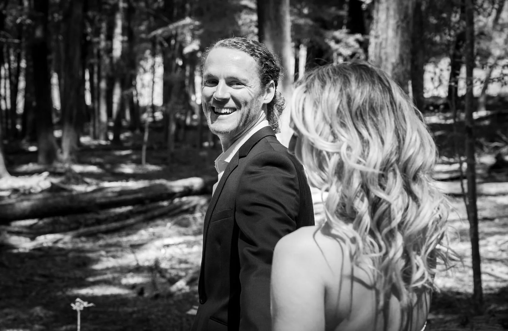 A smiling man in a suit turns to look back at a woman with long, curly hair in an outdoor wooded area.