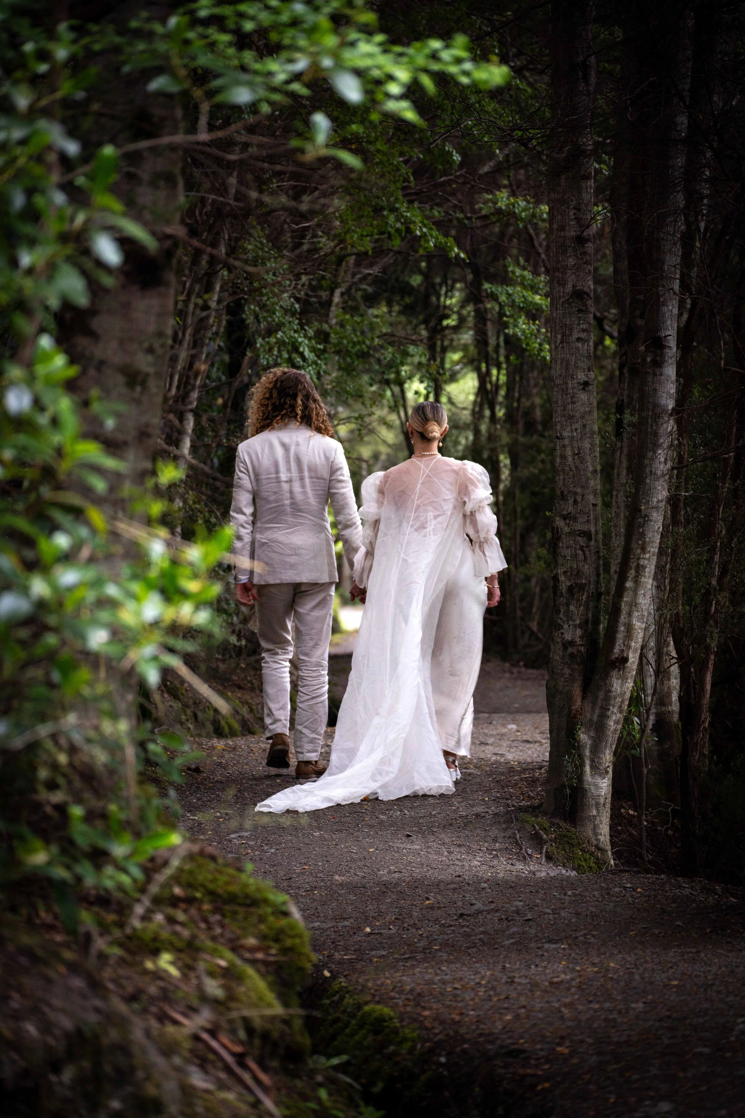 A couple holding hands and walking through a wooded forest, with the woman wearing a white wedding dress and the man in a light-colored suit.