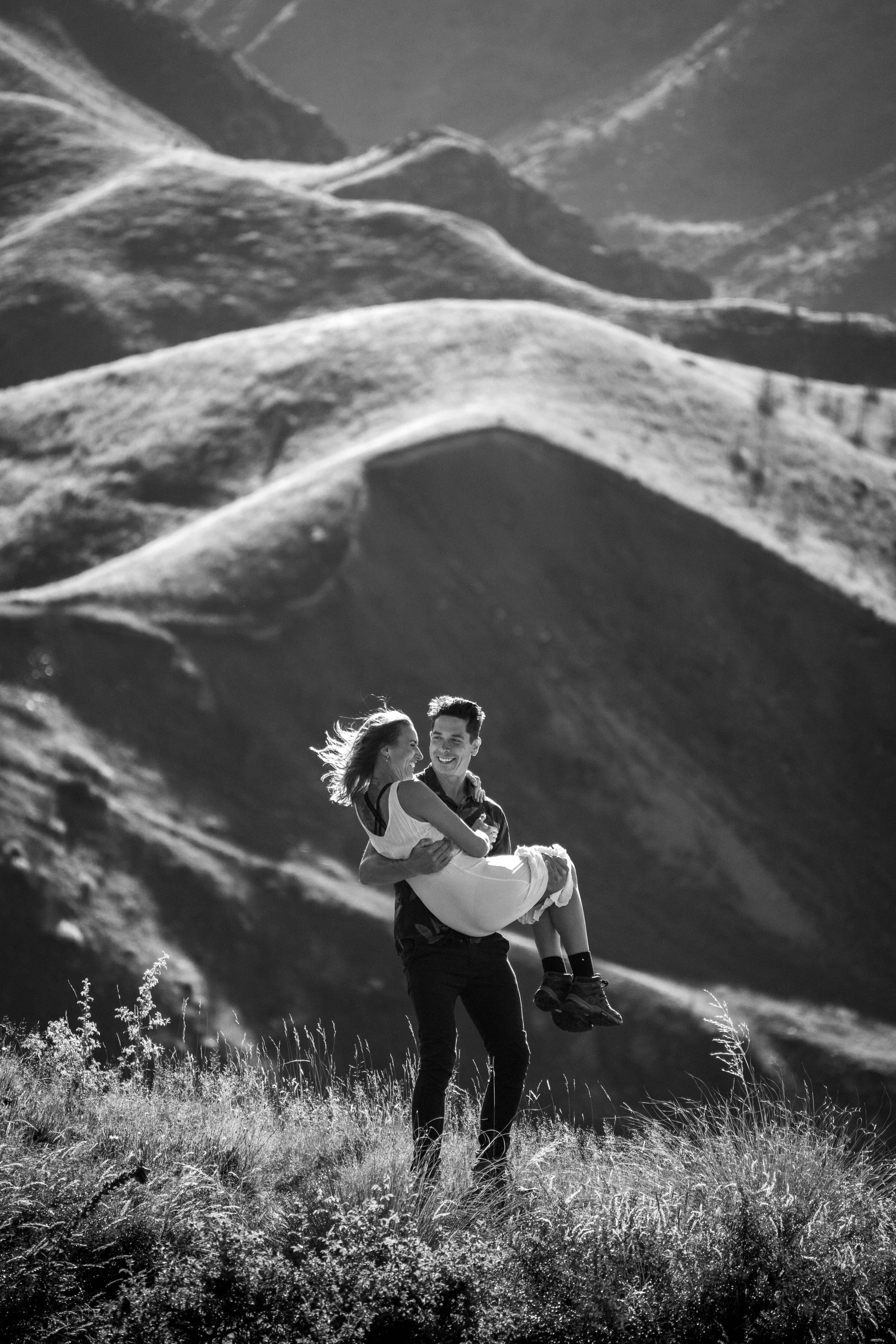 A man lifting a woman in his arms in an outdoor setting with rolling hills and grass, black and white photograph.