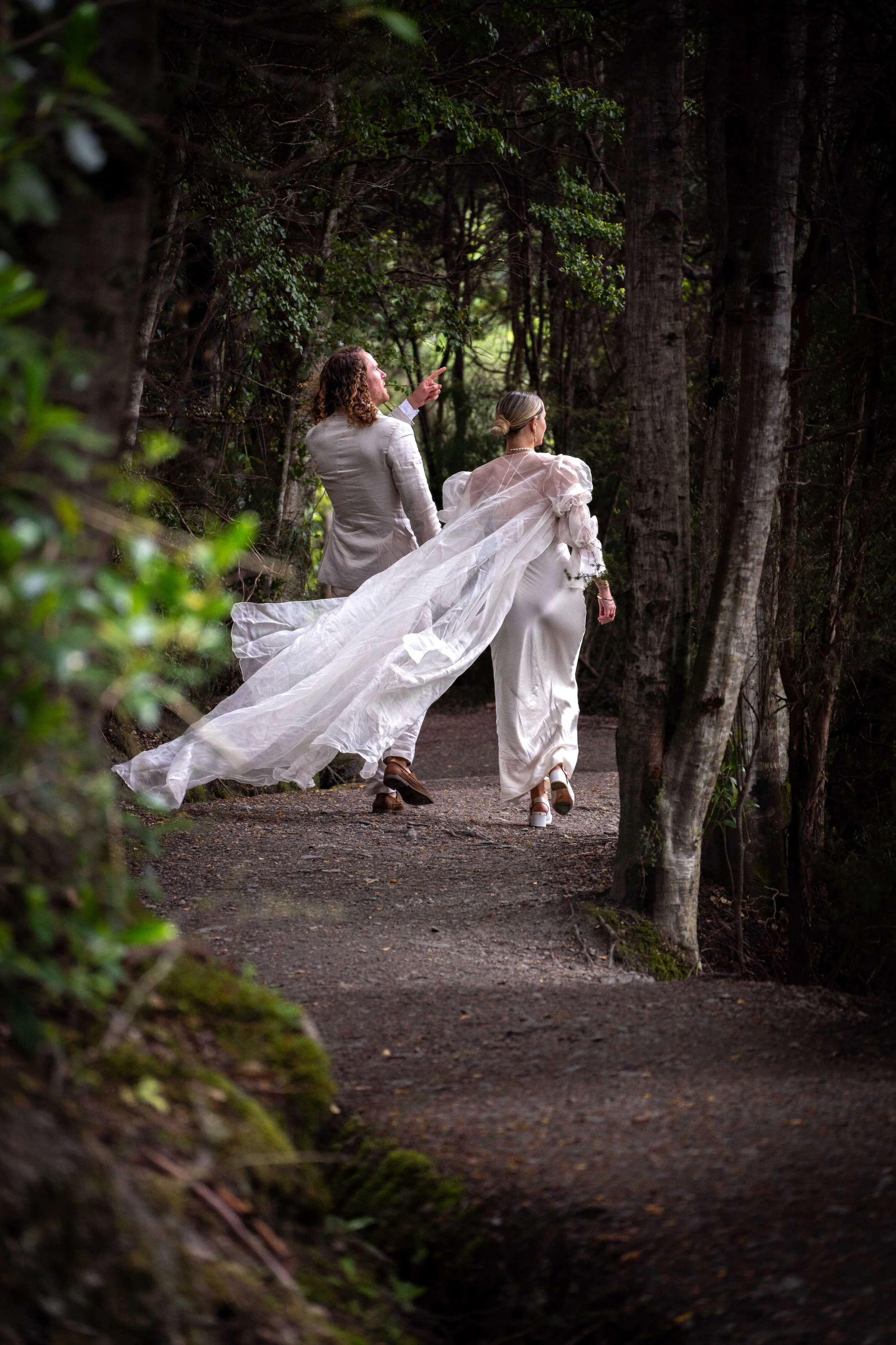 Two women, one in a wedding dress with a flowing veil and another in a suit, walk together along a forest trail surrounded by trees.