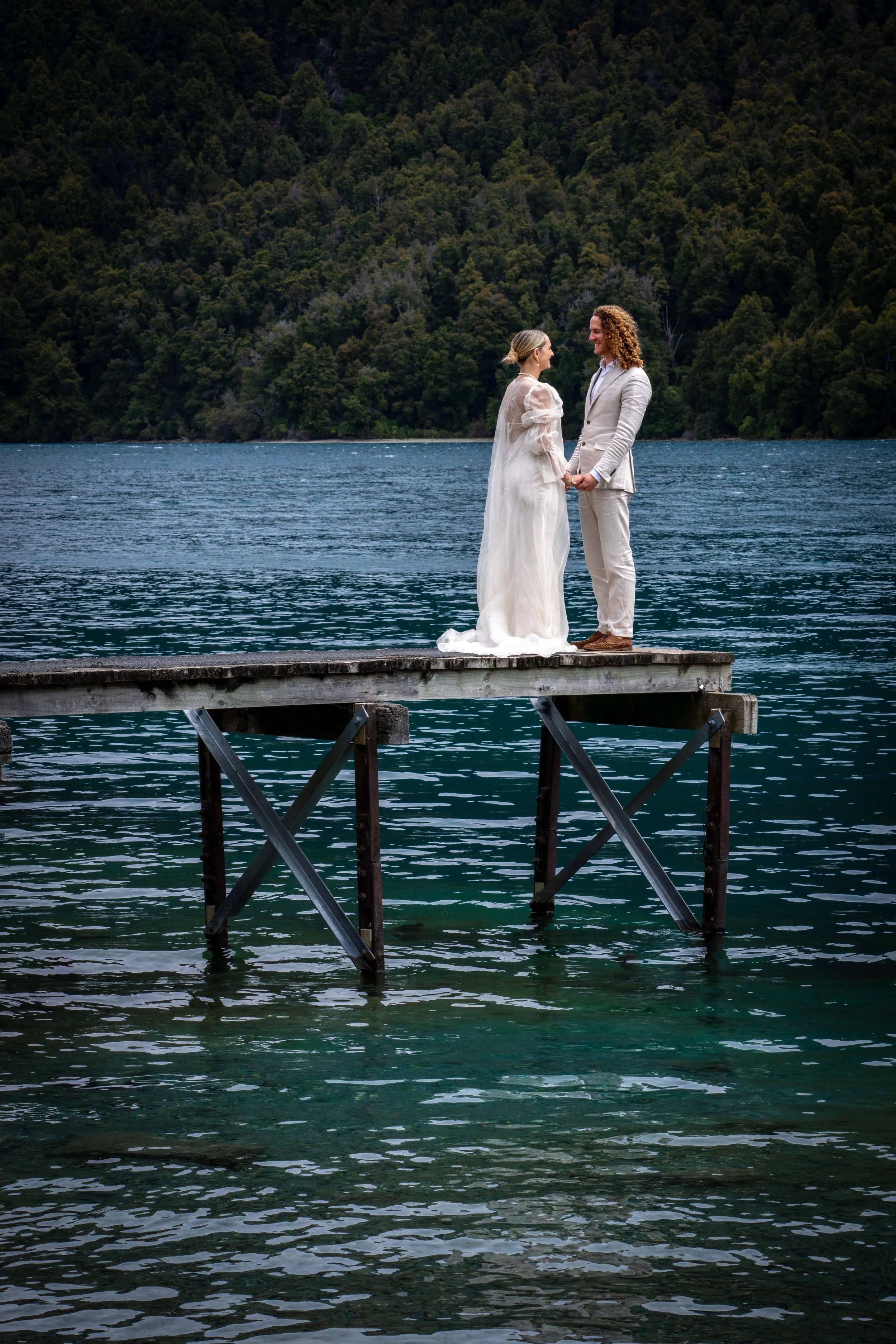 A couple dressed in wedding attire standing on a wooden dock by a lake, holding hands and looking at each other, with forested hills in the background.