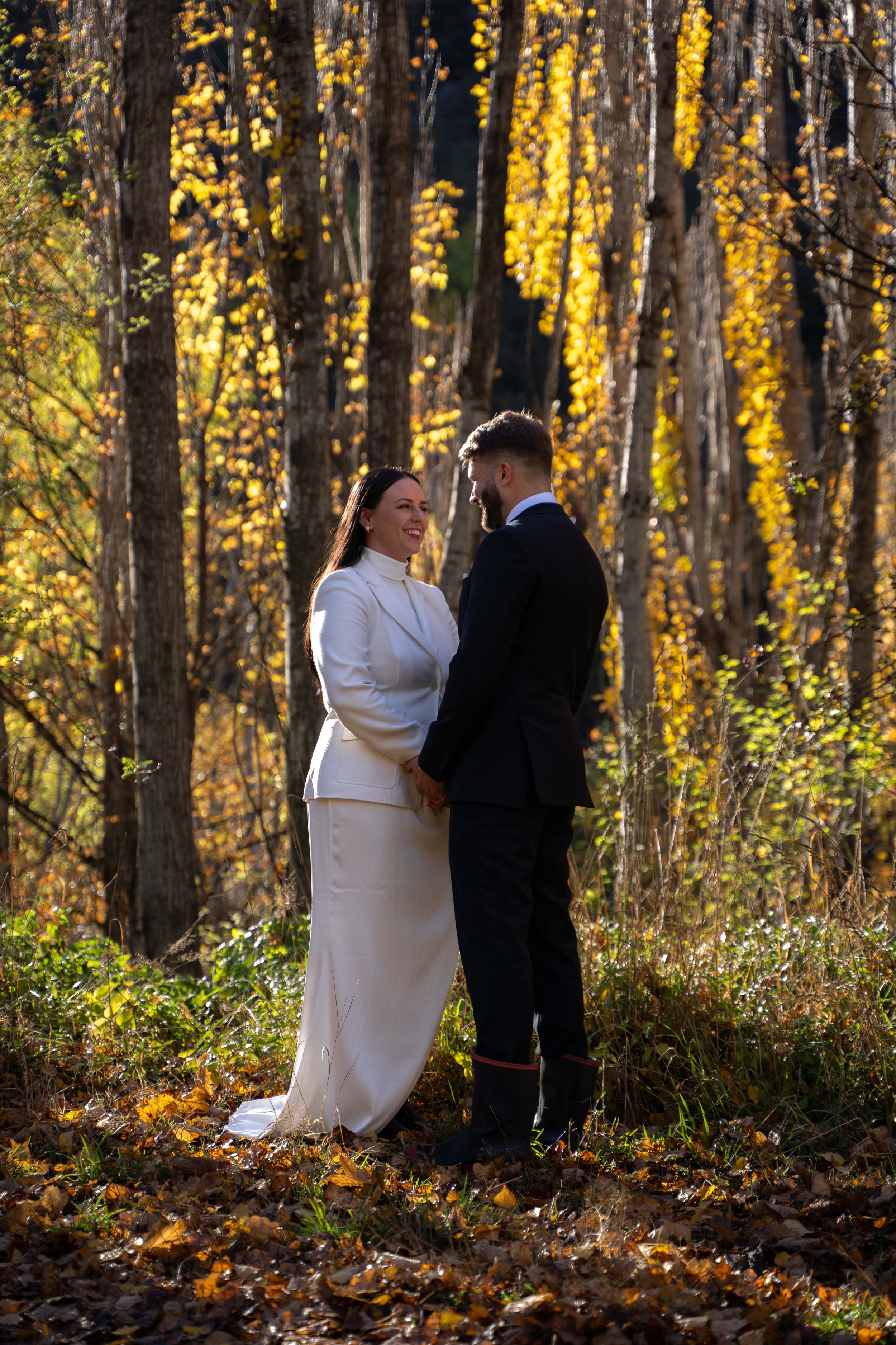 A couple dressed in wedding attire holding hands and smiling at each other in a forest with yellow autumn leaves.