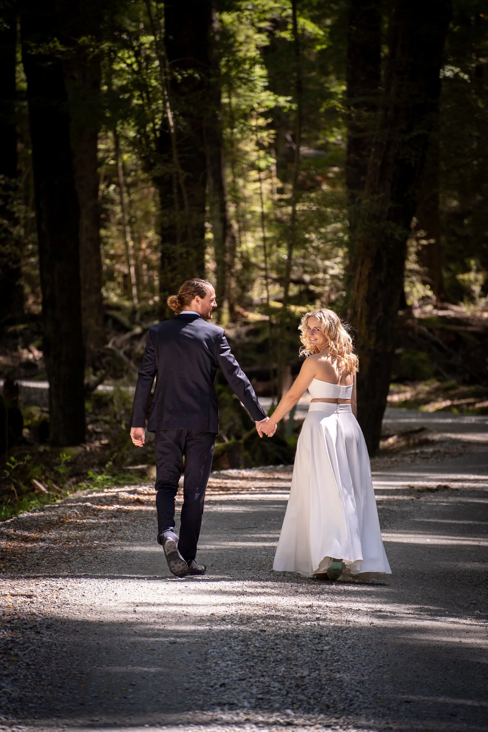 A couple in wedding attire walking hand in hand on a forest trail, surrounded by tall trees, with sunlight filtering through the leaves.