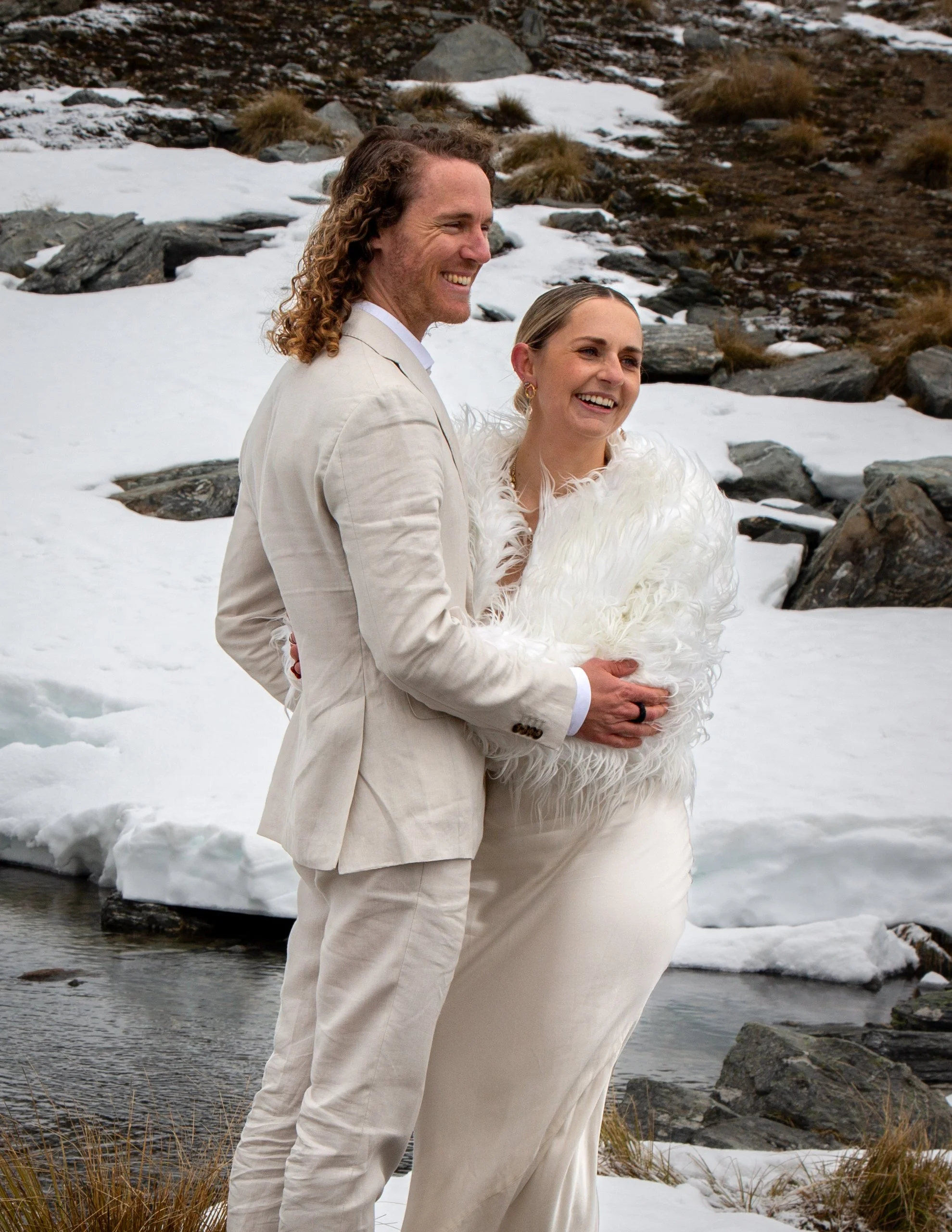 A couple dressed in wedding attire, standing outside near a snow-covered landscape with rocks and a small stream, smiling and holding each other.