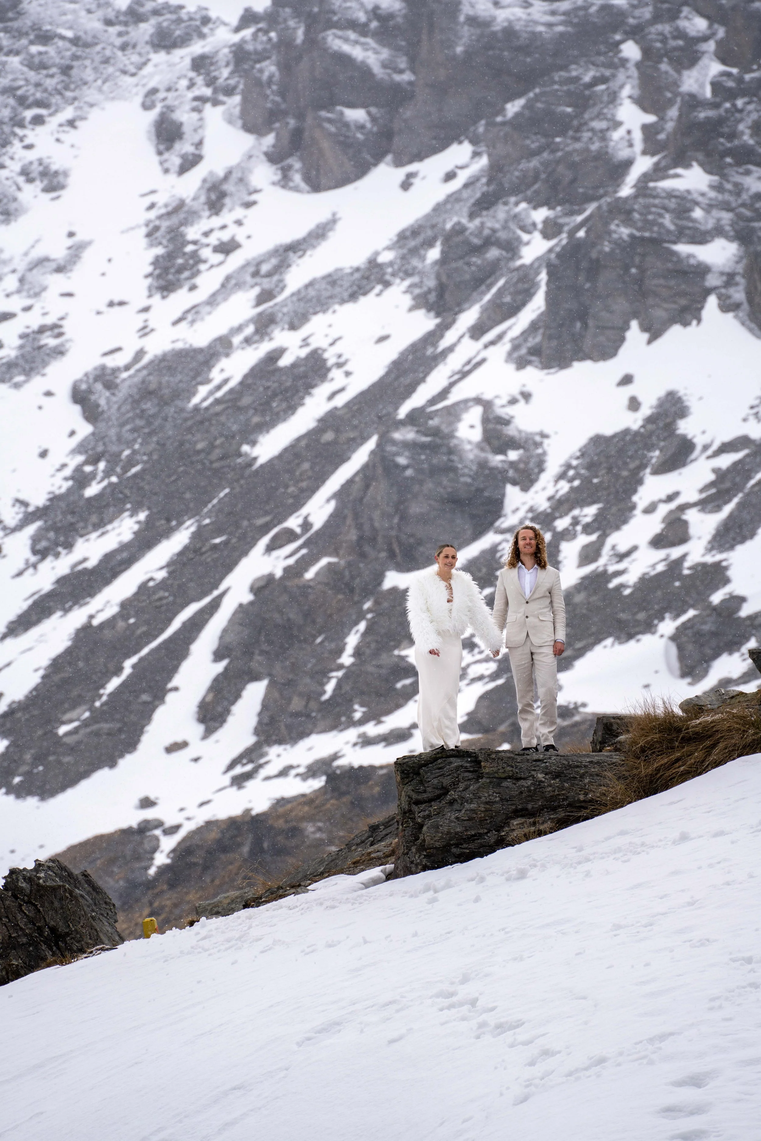 A couple in white wedding attire holding hands and standing on a rock in snowy mountainous terrain.