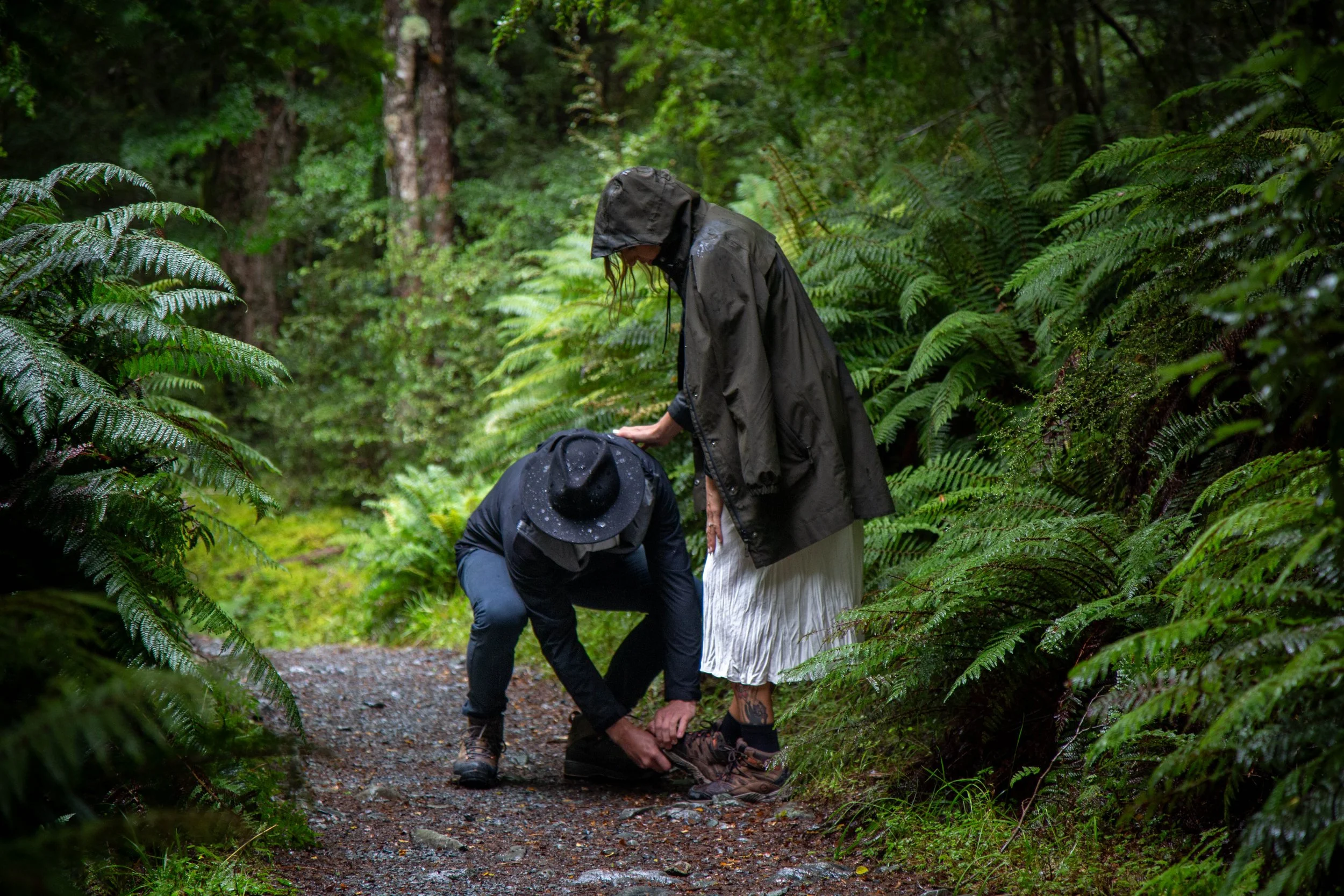 A man crouching down to repair his wife's hiking boots, while she leans on his shoulder, on a forest trail surrounded by lush green ferns and trees.