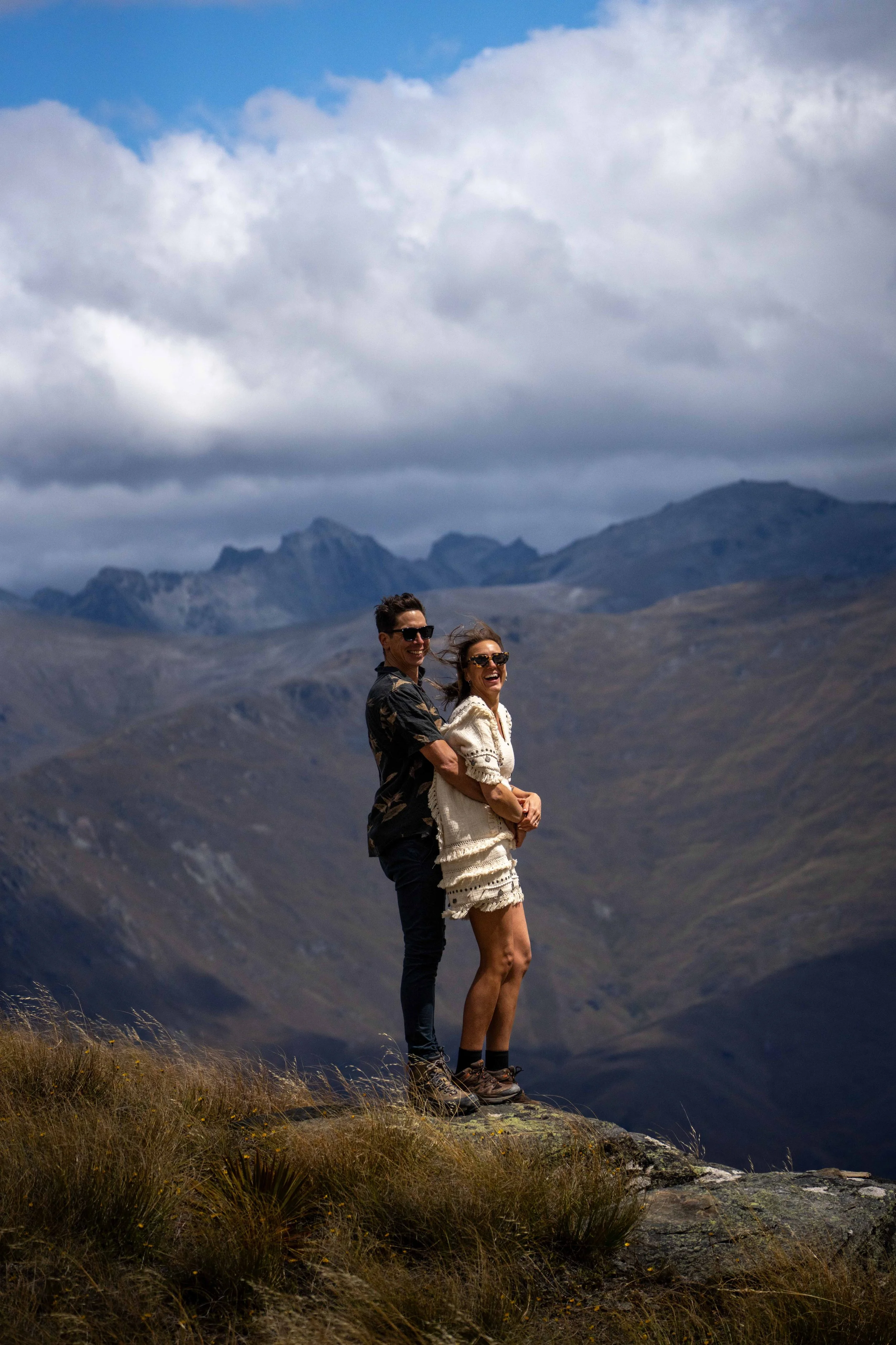 Two people smiling and hugging on a rocky ledge in a mountainous landscape with cloudy skies.