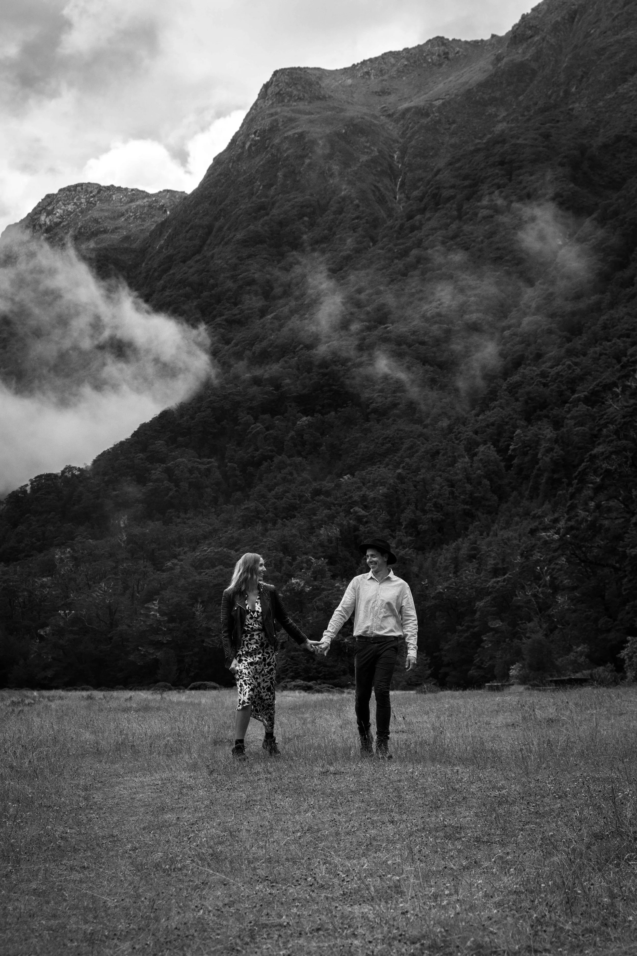 A couple holding hands and smiling while walking across a grassy field with a backdrop of mountains and trees in black and white.