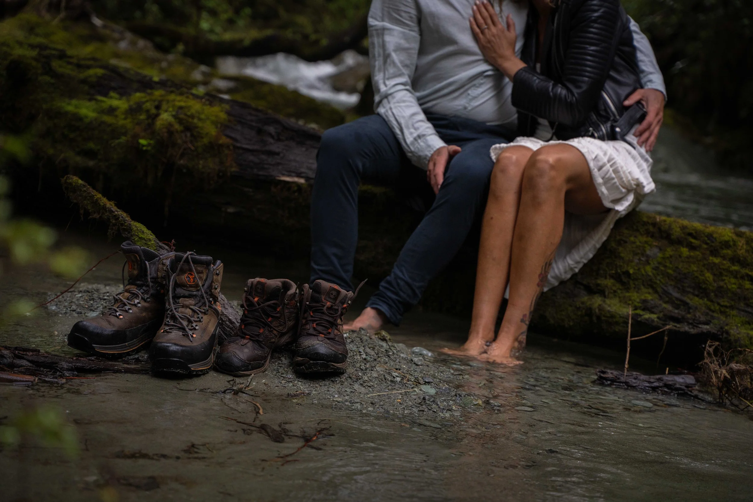 A couple sitting on a mossy log in a creek, with their feet in the water. Three pairs of hiking boots are on the ground nearby.