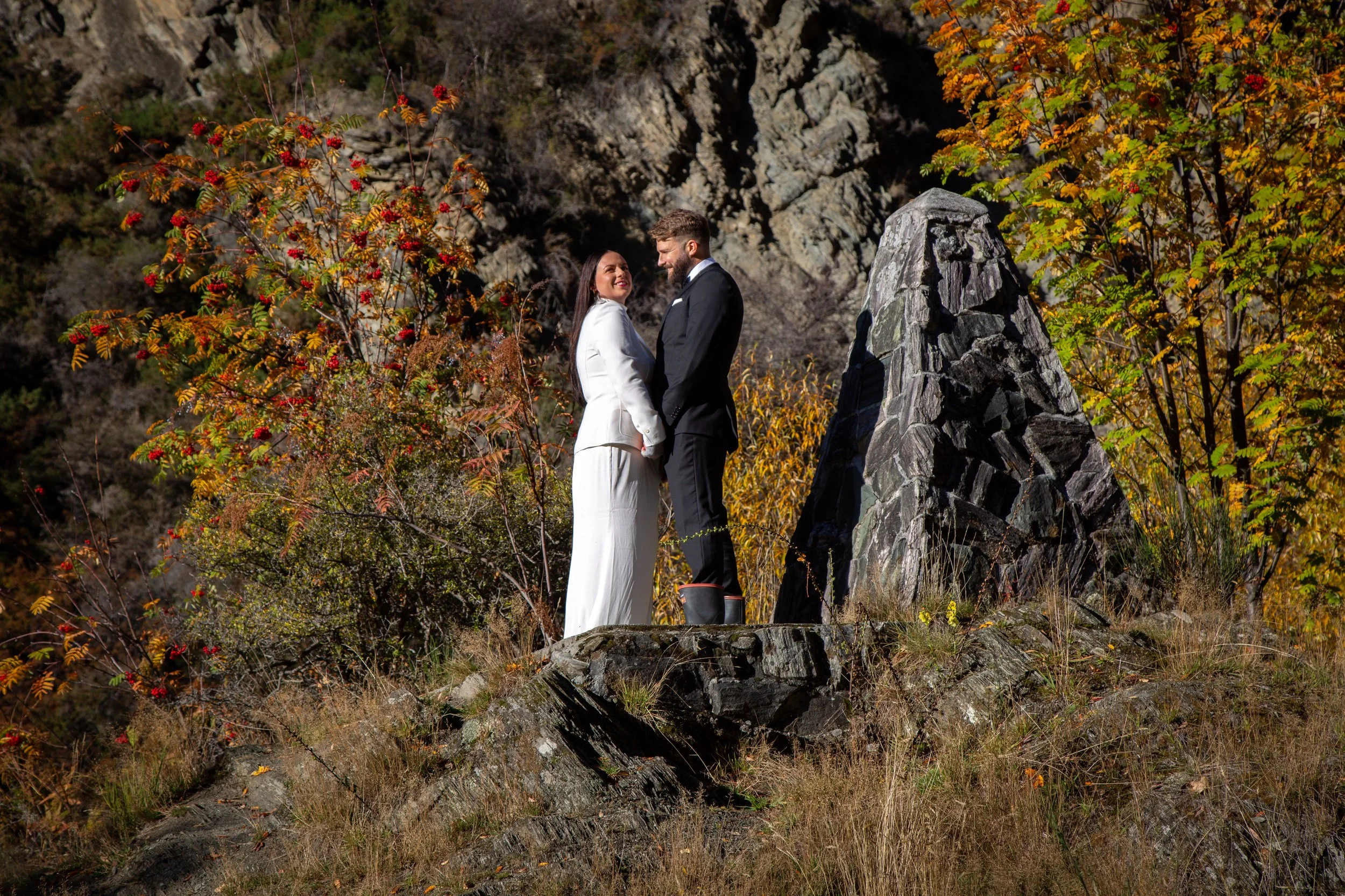 A couple dressed in wedding attire standing on rocks outdoors during fall. The woman wears a white suit and the man in a black suit, holding hands and smiling at each other. There is colorful foliage and a large rock formation in the background.