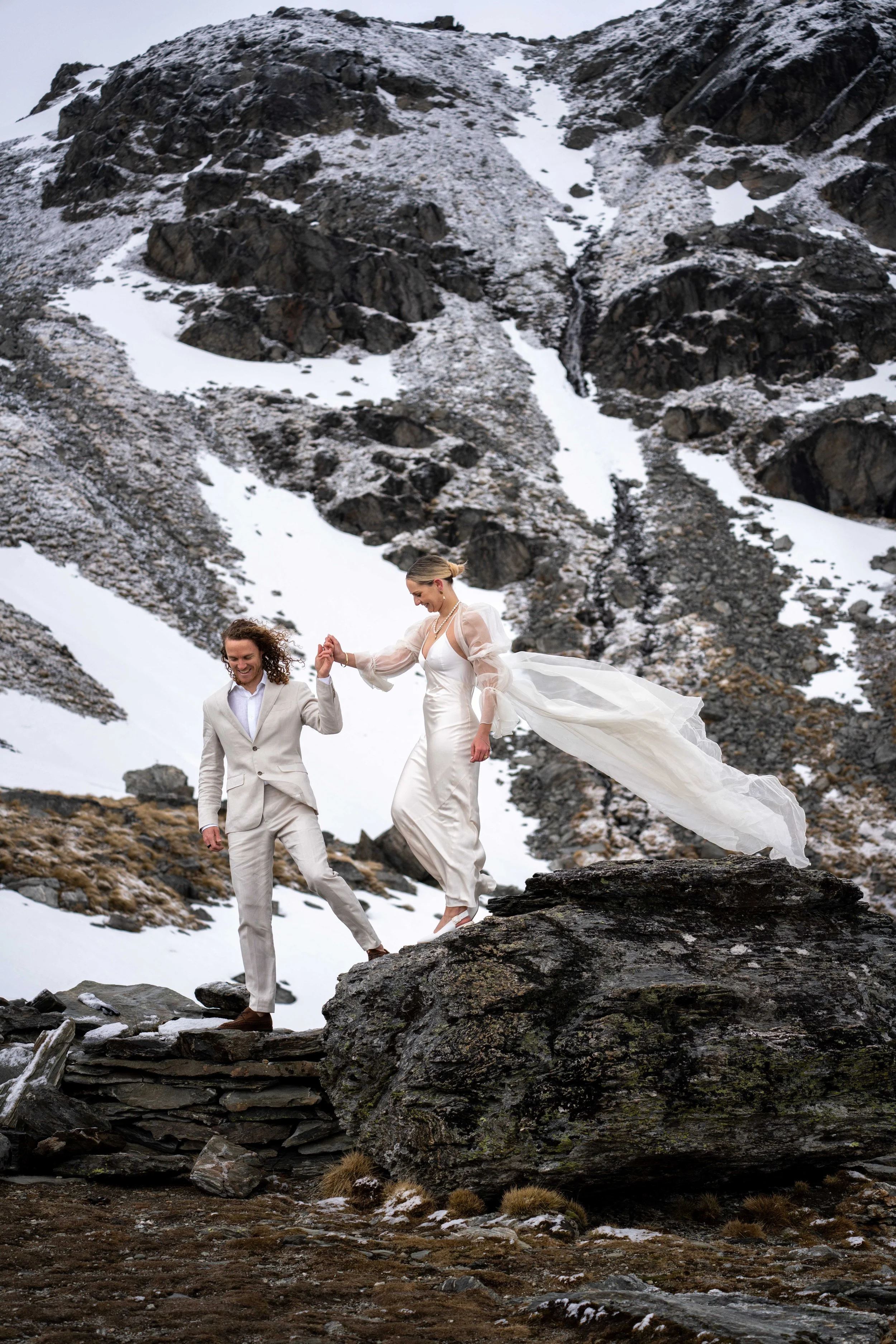 A couple dressed in wedding attire holding hands and balancing on a large rock in a snowy mountain landscape.
