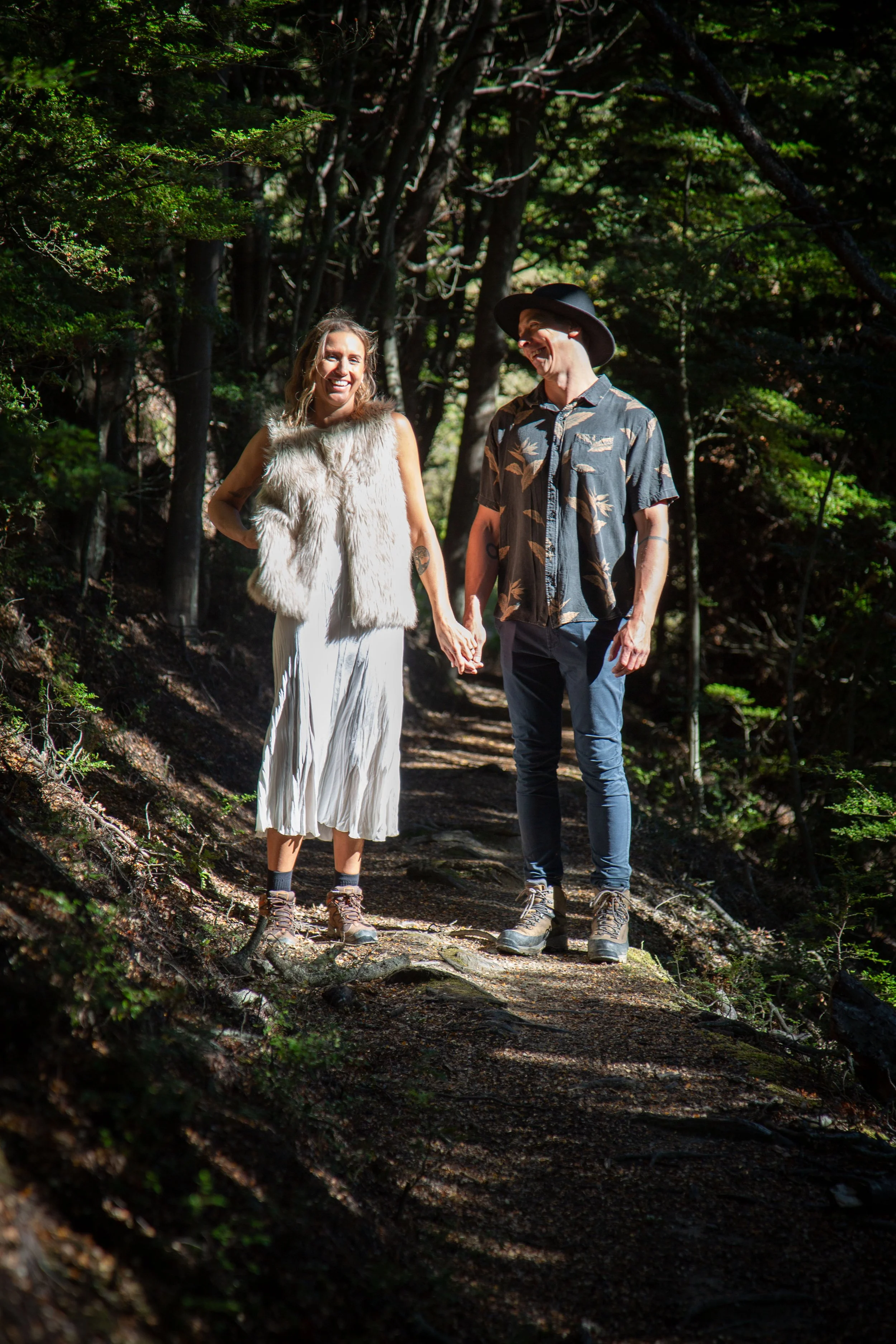 A man and woman holding hands and smiling while walking on a forest trail surrounded by trees.