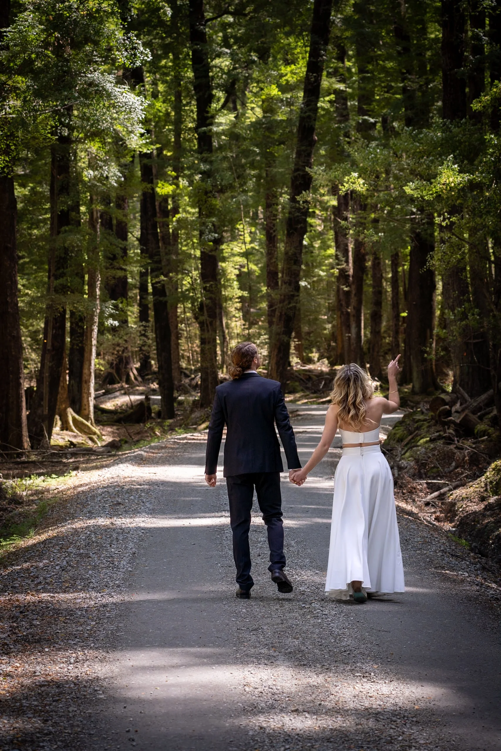 A couple dressed in wedding attire walking hand in hand down a forested dirt path, with the woman pointing up.