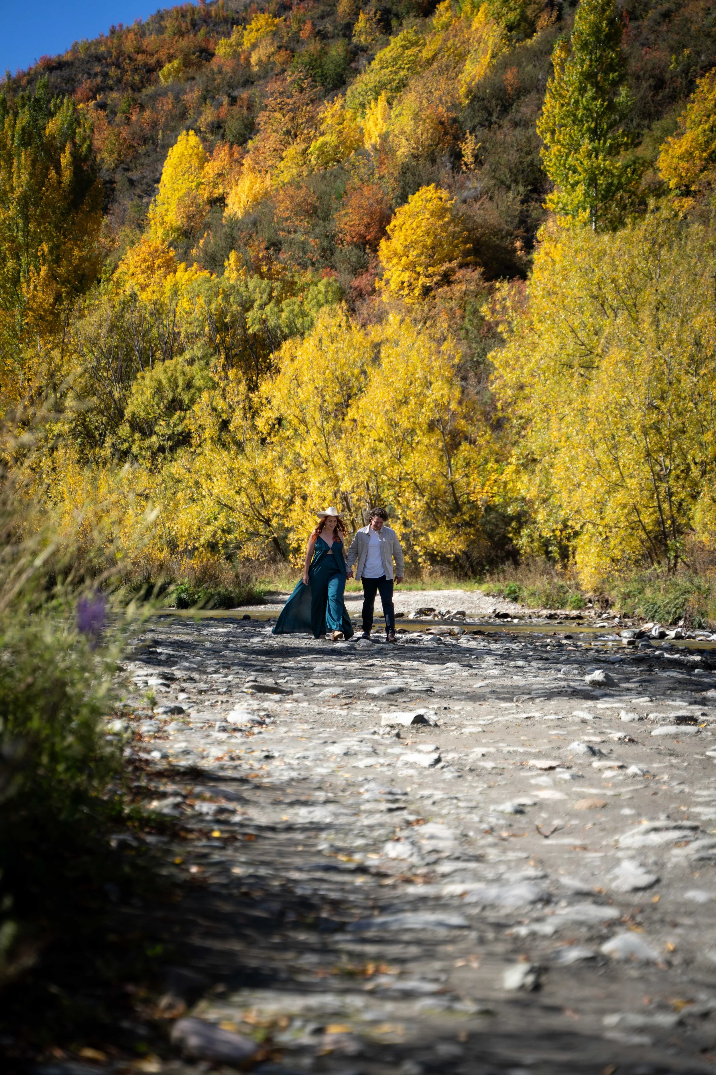 A couple walking together along a rocky riverbed surrounded by fall foliage on a hillside.