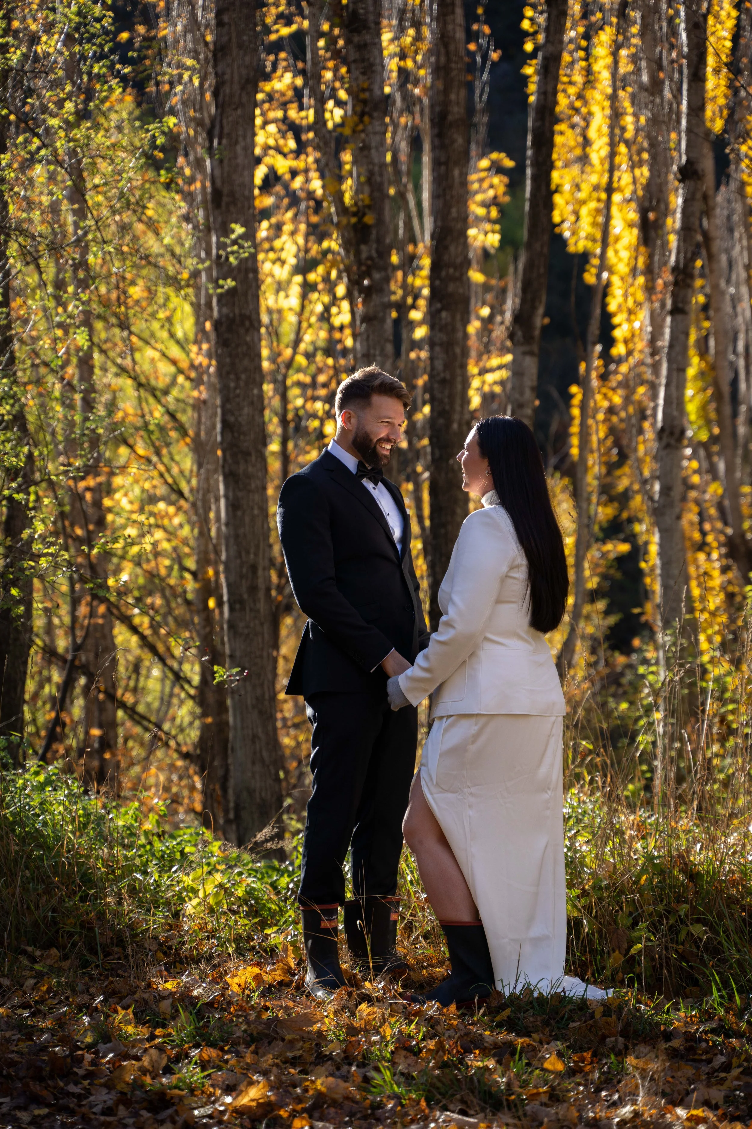A couple dressed in wedding attire standing in a wooded area with autumn leaves, holding hands and smiling at each other.