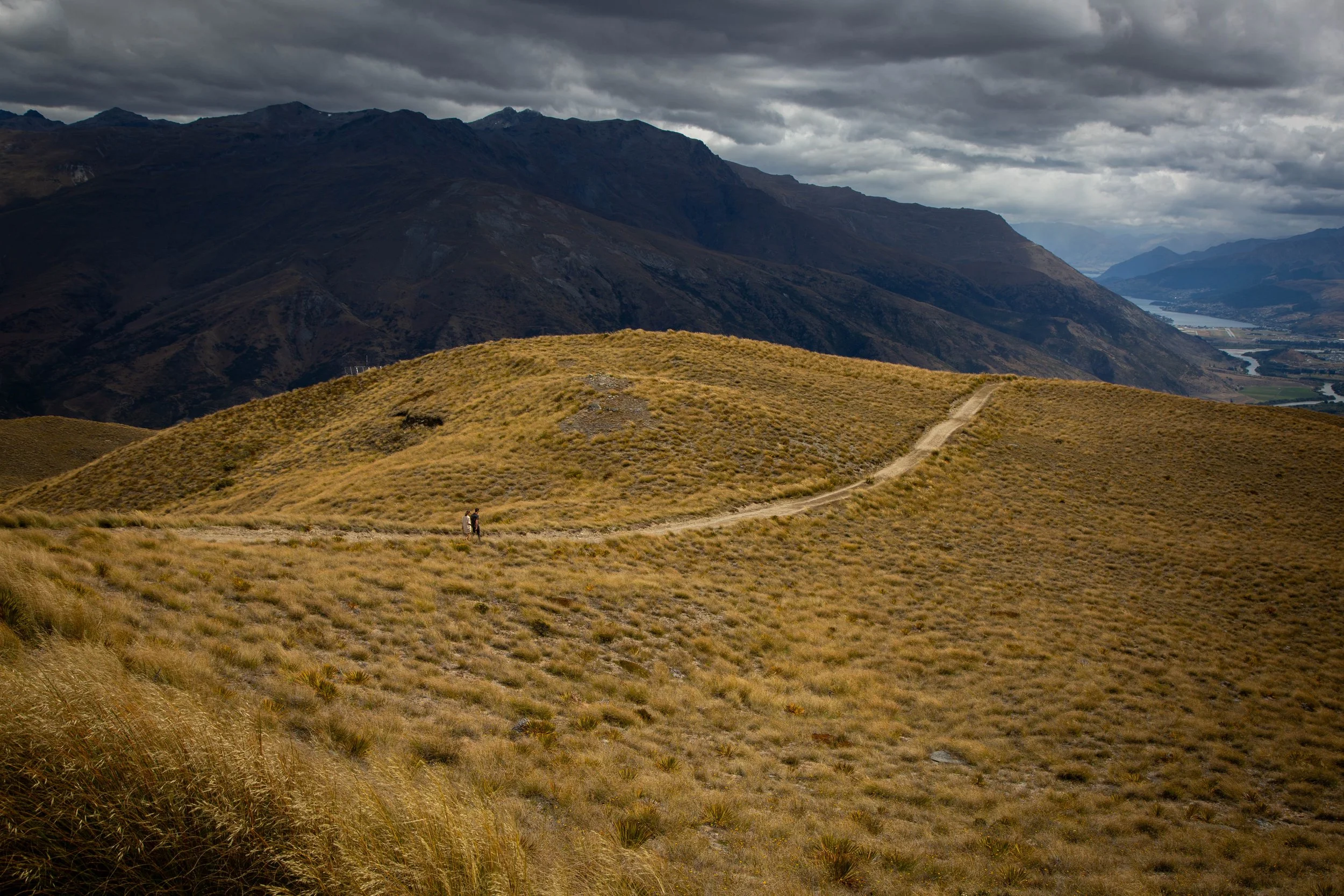 Hiking trail winding through grassy hills with a mountain range and cloudy sky in the background, and a tiny couple walking the trail in the distance.