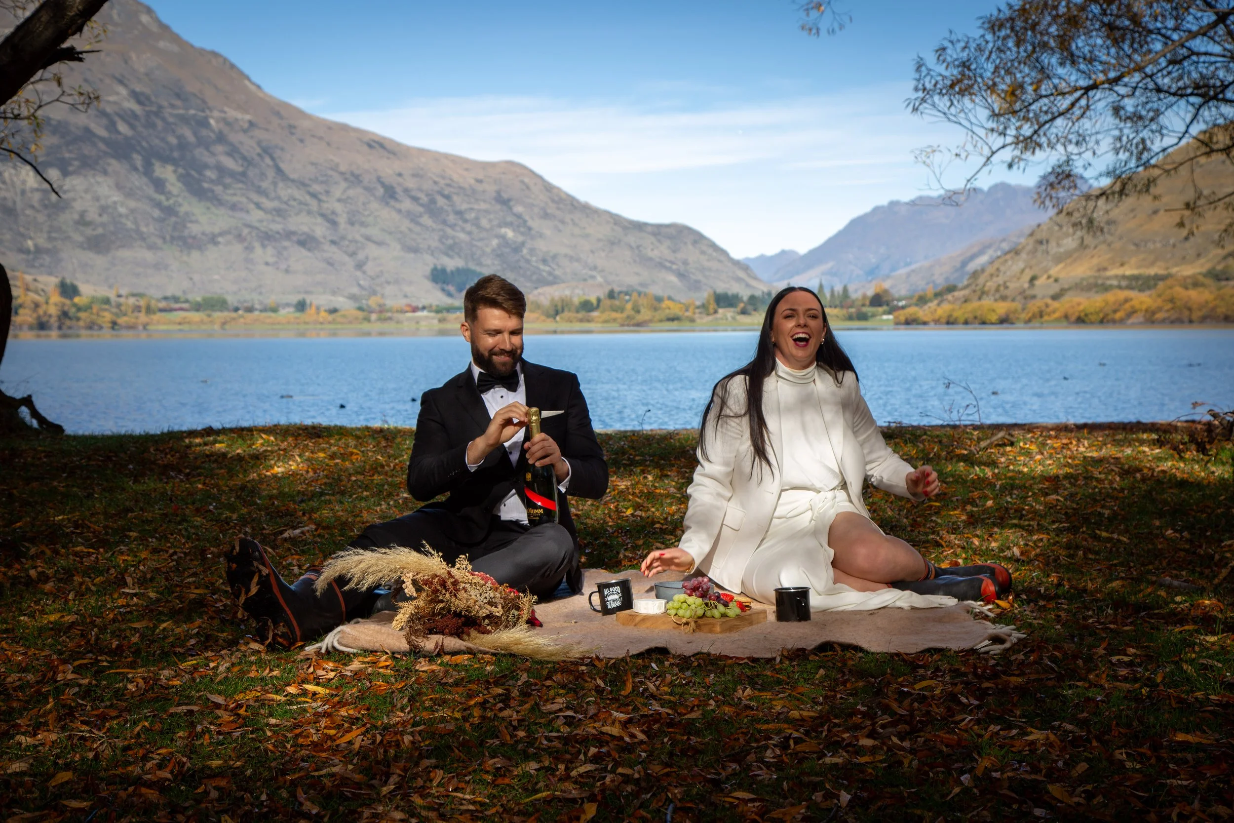 A man and woman having a picnic on a blanket by a lake with mountains in the background. The man is opening a bottle of champagne, and the woman is laughing. There are grapes and mugs on the blanket.