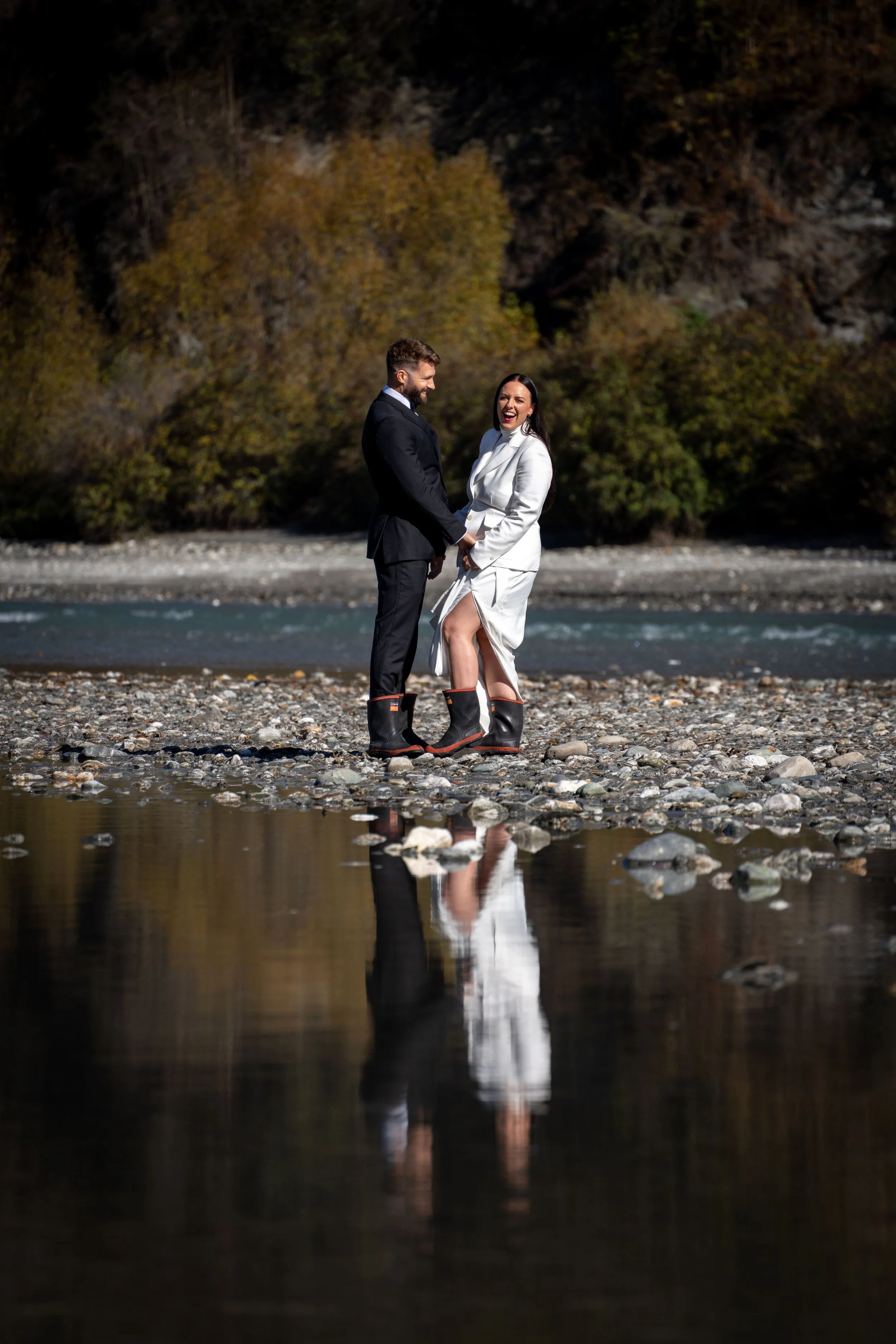 A couple in formal attire standing on a rocky riverbank, holding hands and smiling at each other. The woman is wearing a white dress with black boots, and the man is in a dark suit. Their reflection is visible in the water, with a background of trees and hills.