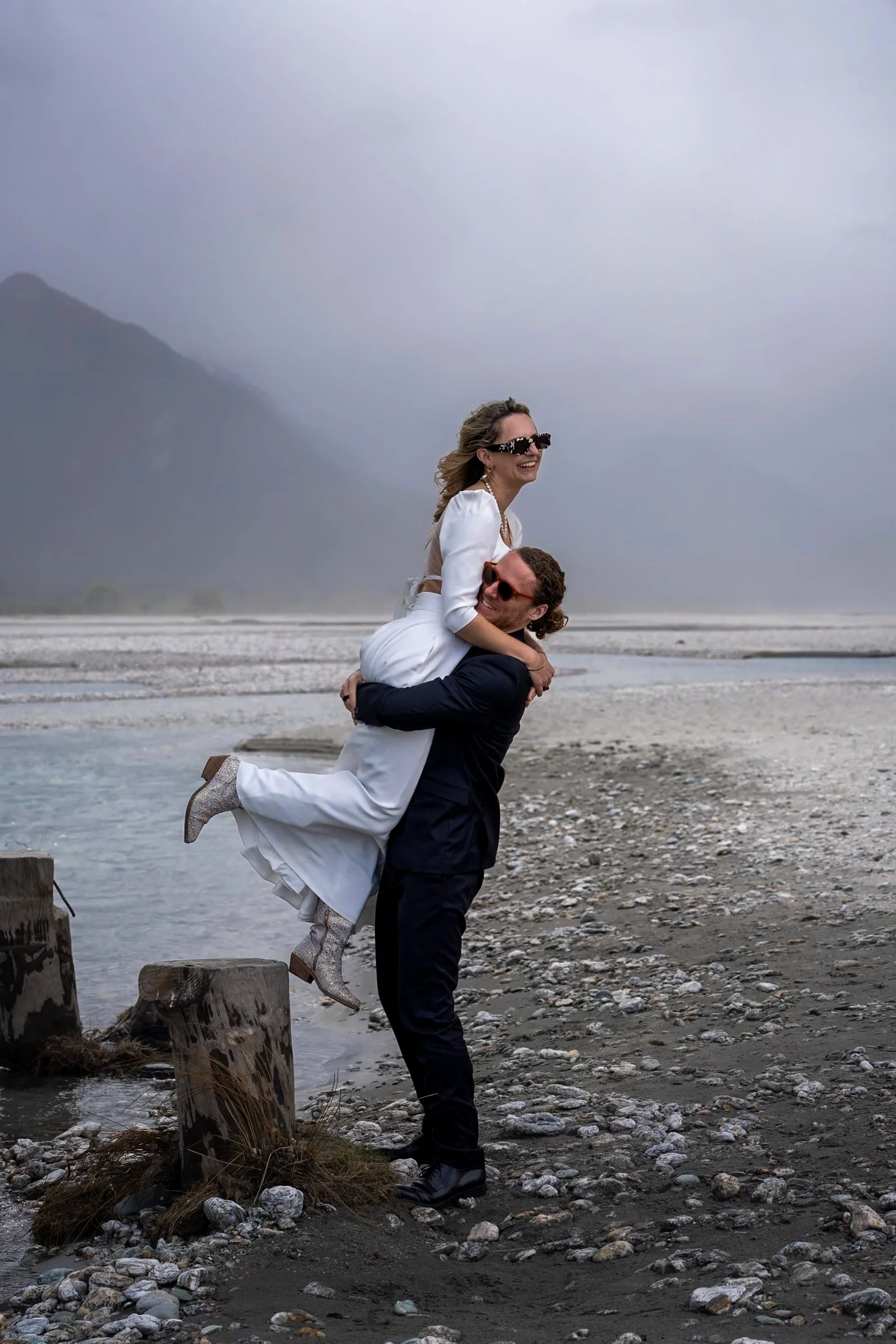 A woman in a white dress and boots is being lifted by a man in a black suit on a rocky beach with a misty mountain background.